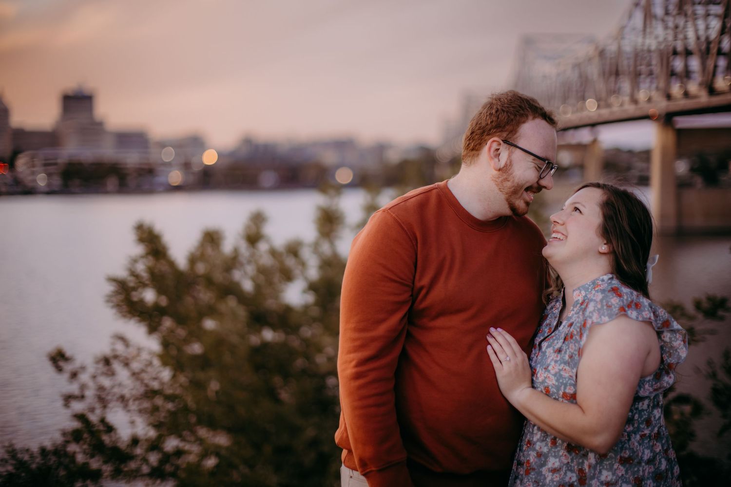 Sweet couple embracing during golden hour by the river with urban backdrop.