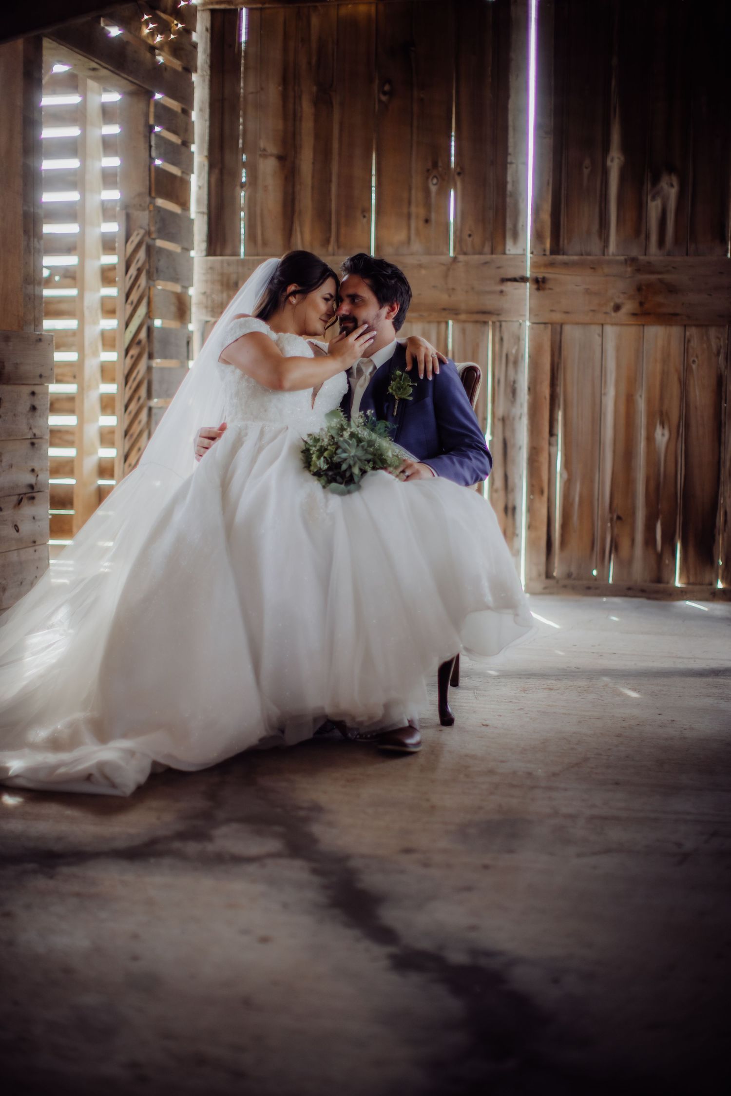Wedding couple in elegant attire pose intimately in a rustic barn with wooden walls.
