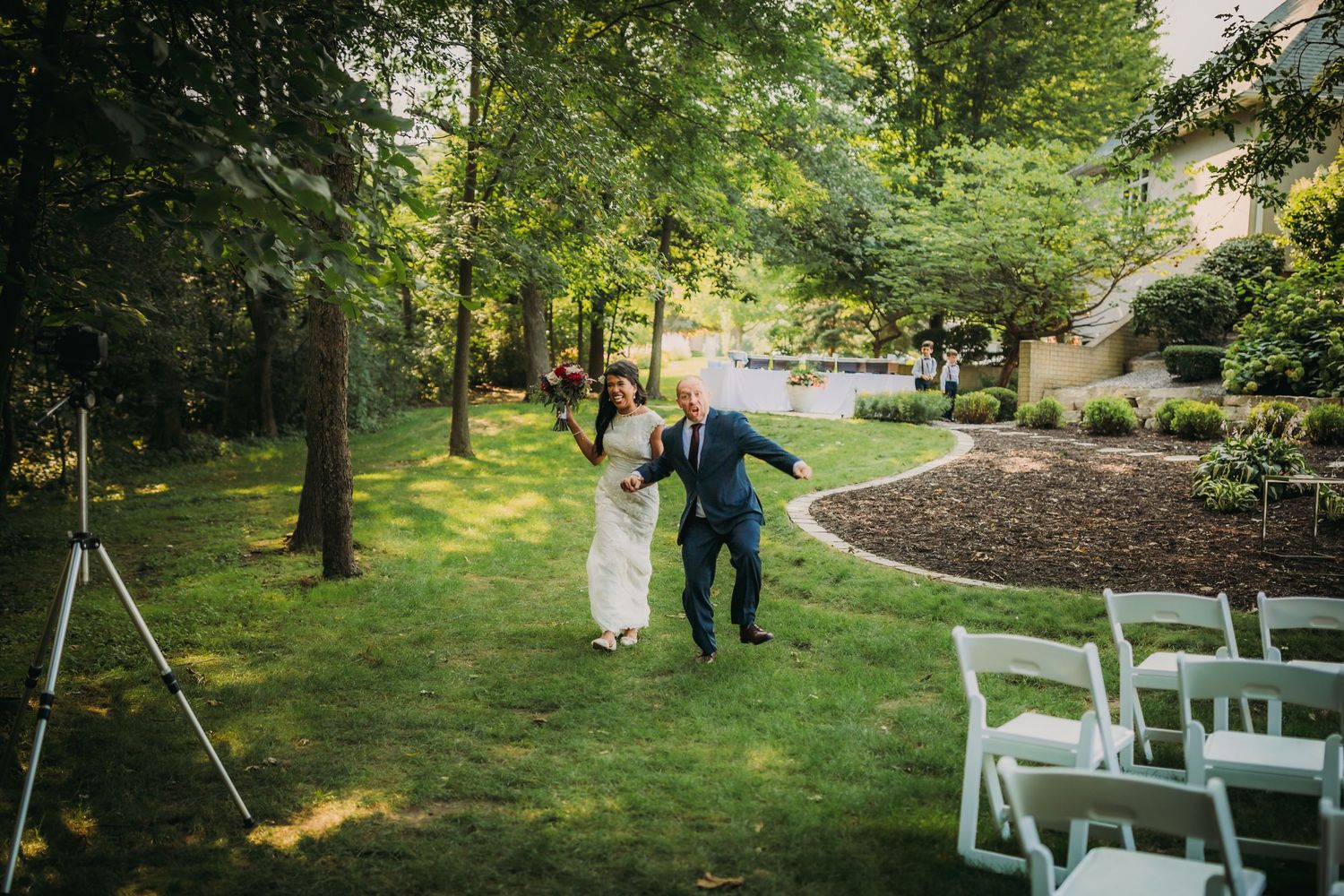 Wedding couple walks down a garden path with white chairs and natural landscaping.