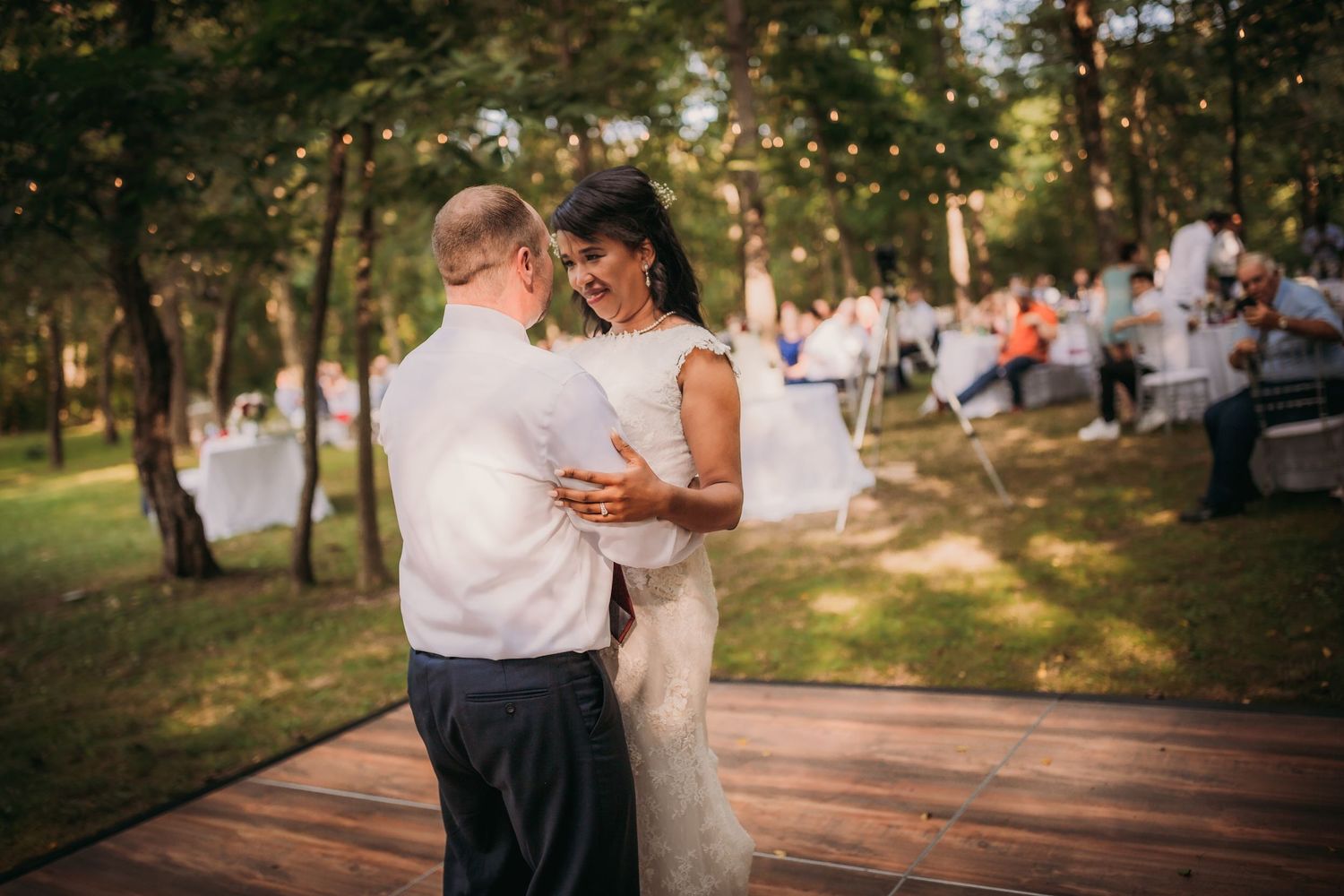A couple embraces during their wedding dance on a wooden platform with guests seated in the background.