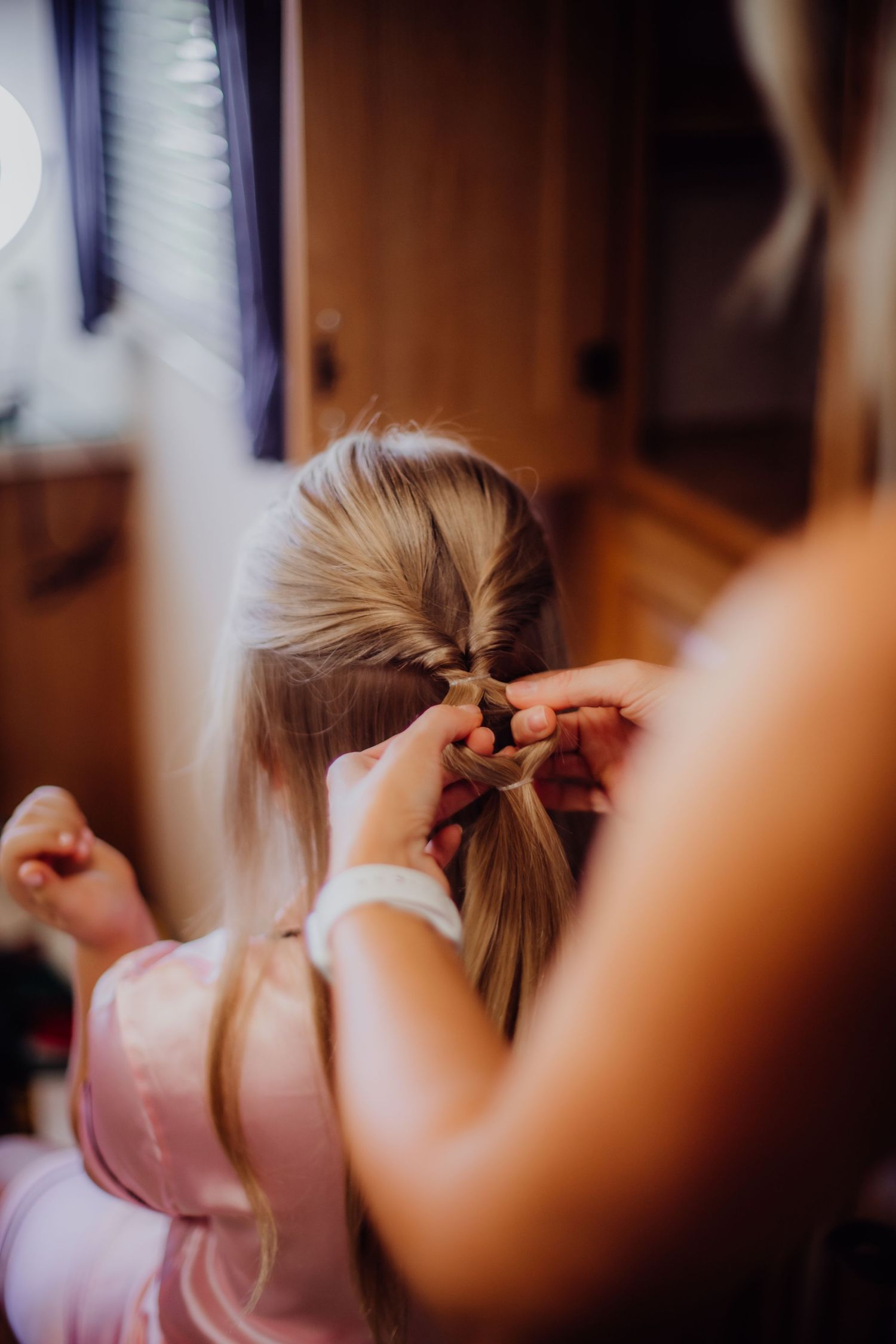 Hands carefully style blonde hair into a neat braid against natural window light.