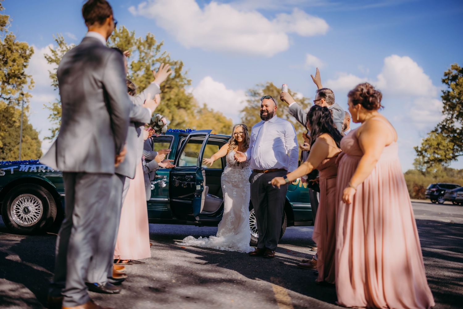 Wedding party members interact beside classic car during outdoor celebration.