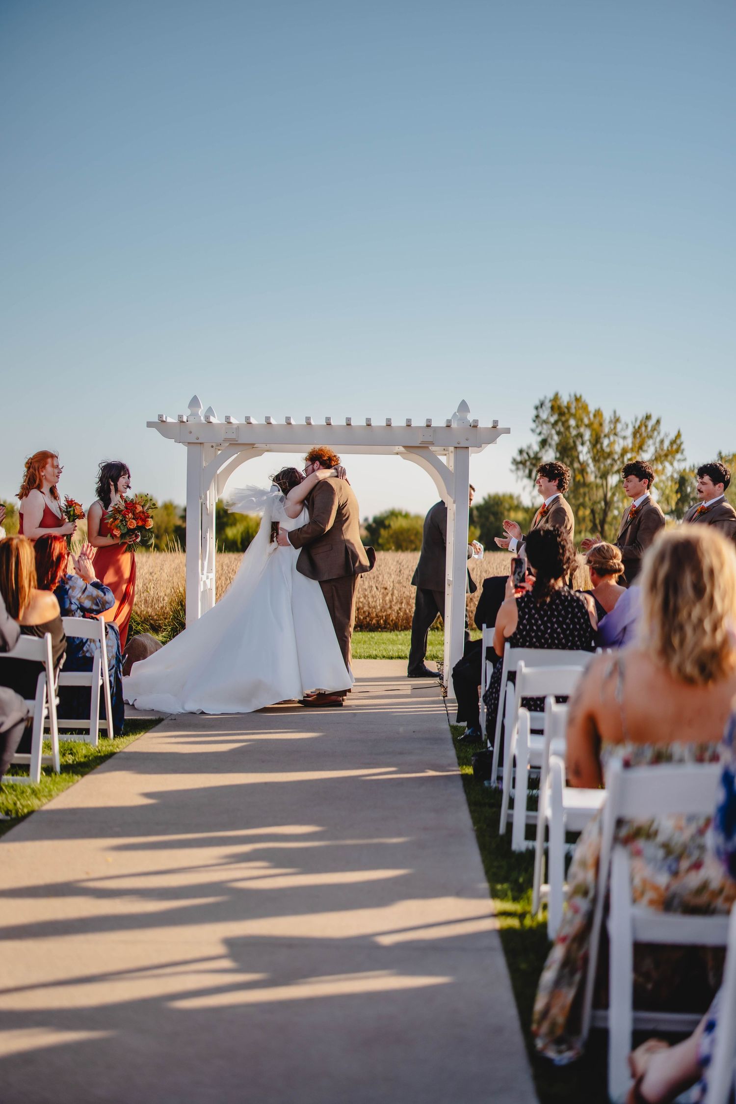 Intimate wedding moment beneath pergola during golden hour with seated guests.