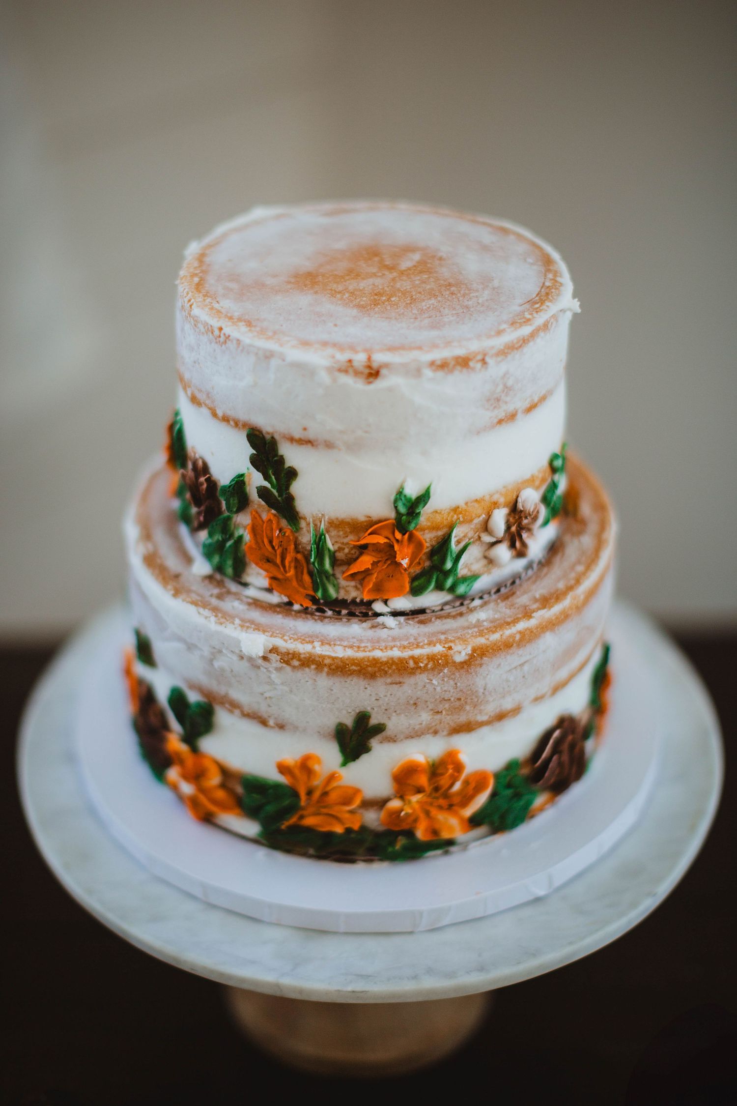 Two-tiered white wedding cake decorated with orange floral designs and greenery on a cake stand.