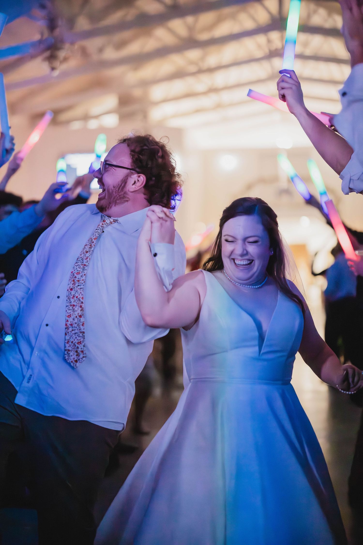 Happy couple dancing enthusiastically at their reception while guests wave light sticks in the air.