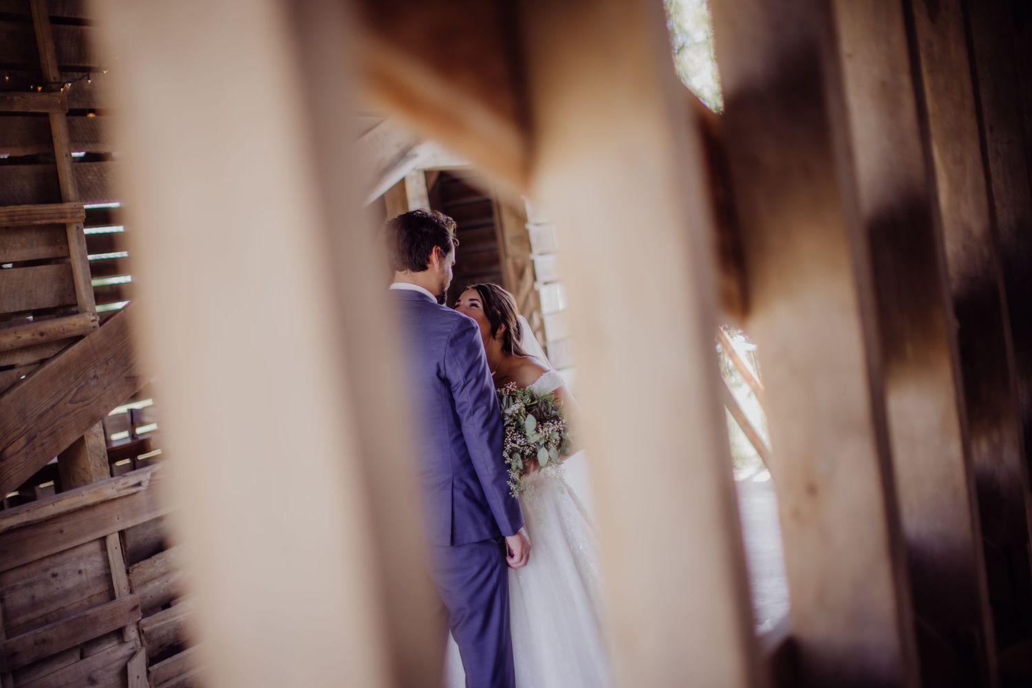Artistic wedding photo through wooden slats showing couple in rustic barn setting.