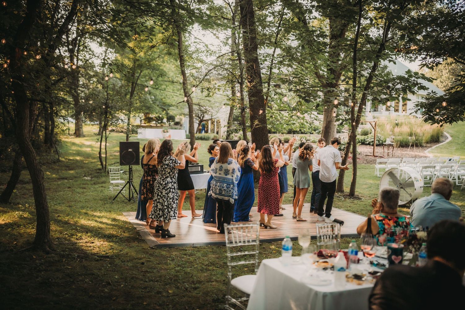 Wedding celebration in a wooded setting with guests dancing and tables decorated for reception.