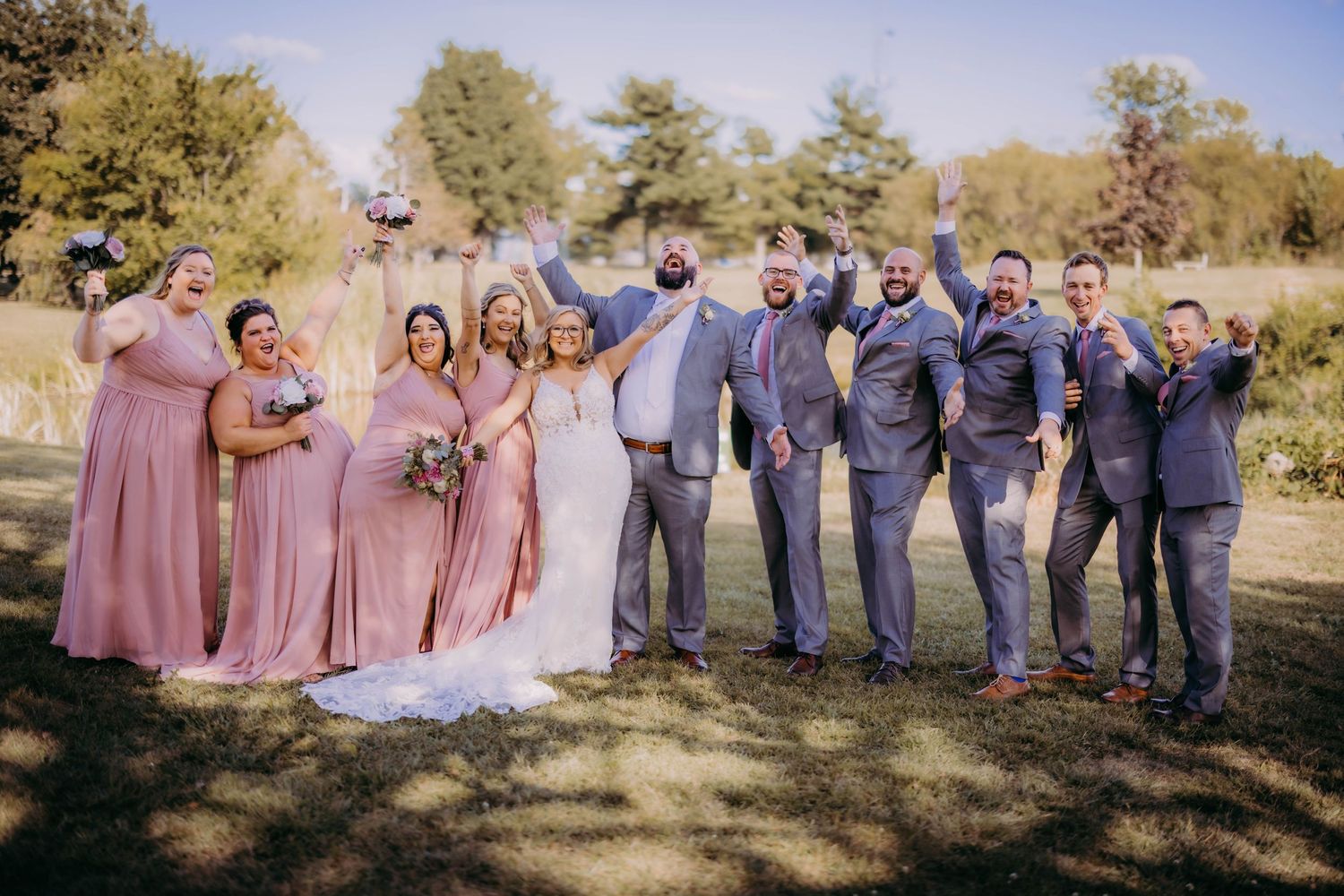 Wedding party in pink dresses and gray suits poses together on sunny autumn day.