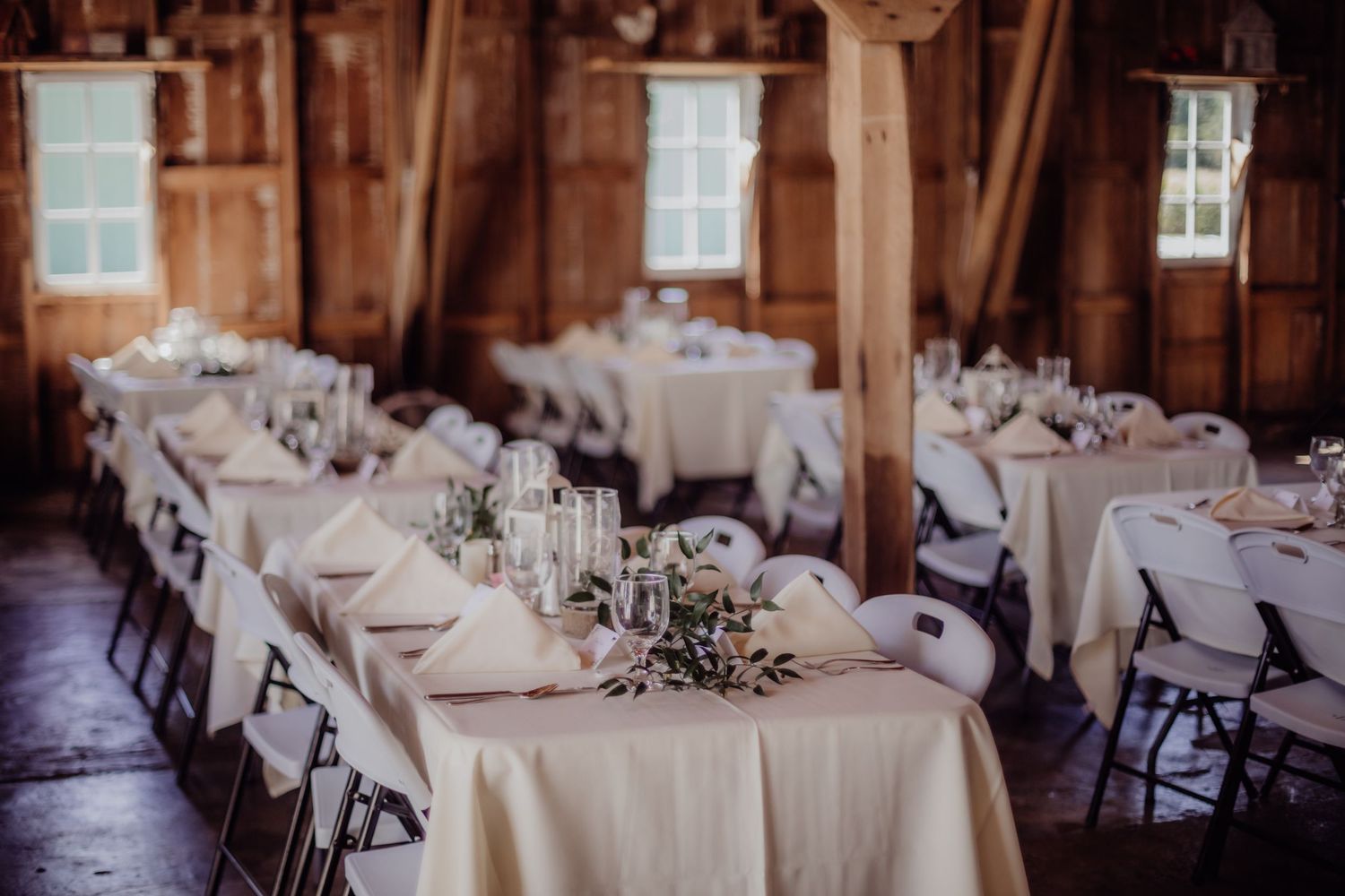 Reception tables set in a rustic barn venue with white linens and natural lighting.