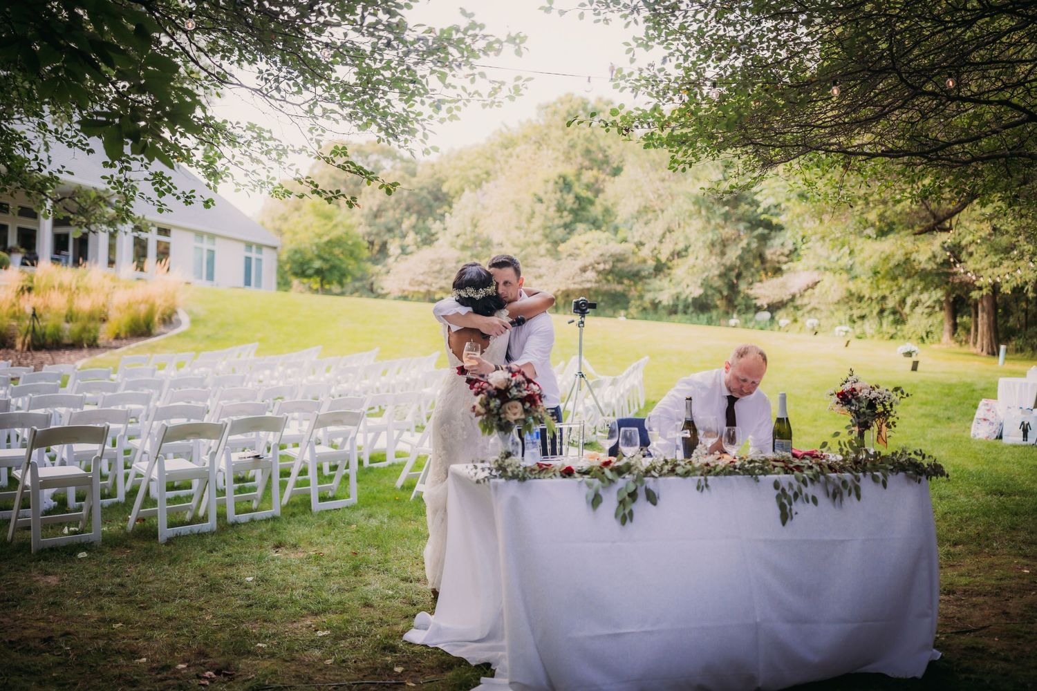 Rustic outdoor wedding venue with white-clothed tables and chairs arranged under tree canopy.