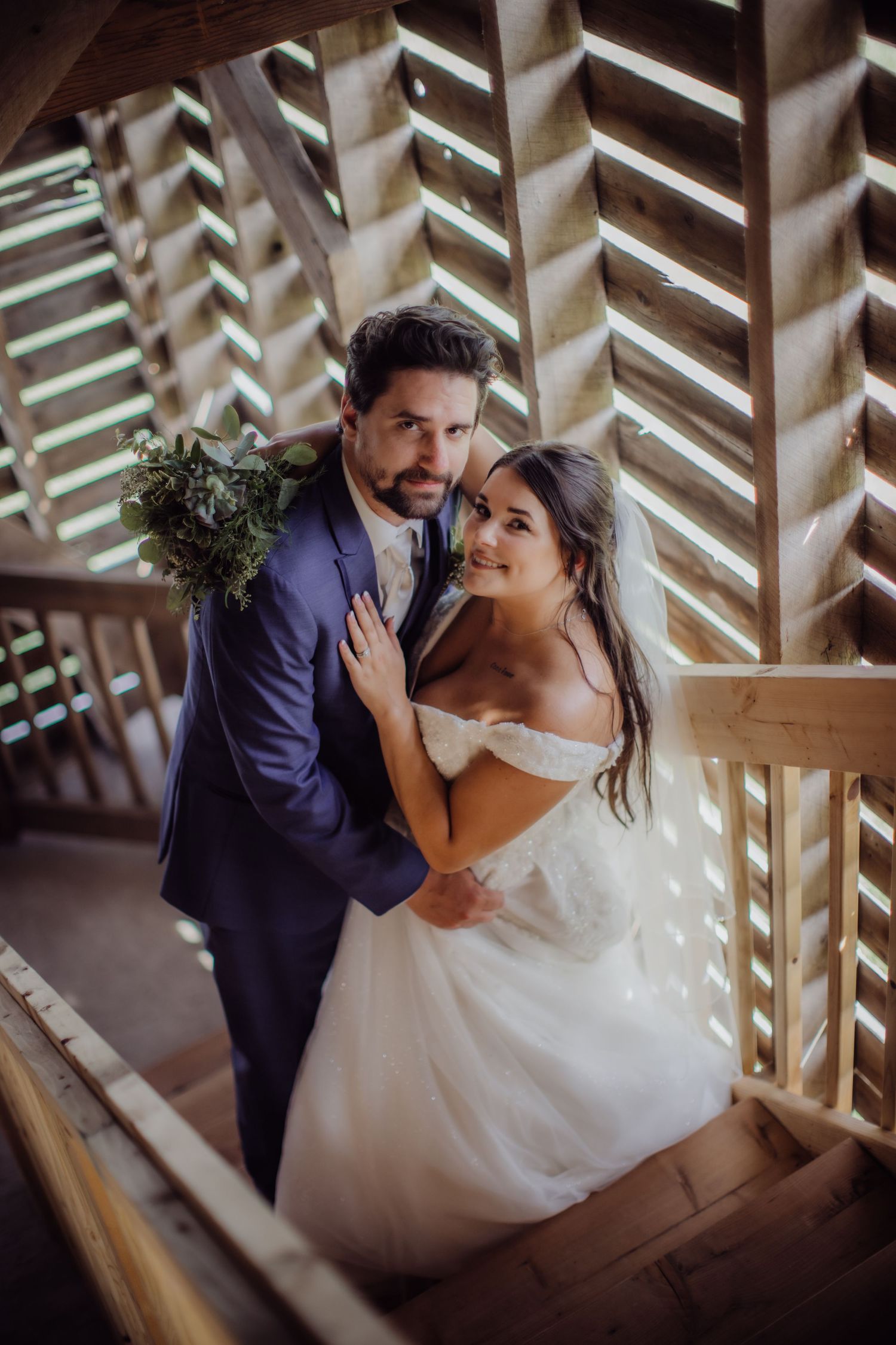 Newlyweds share tender moment on wooden staircase with dramatic architectural lighting.