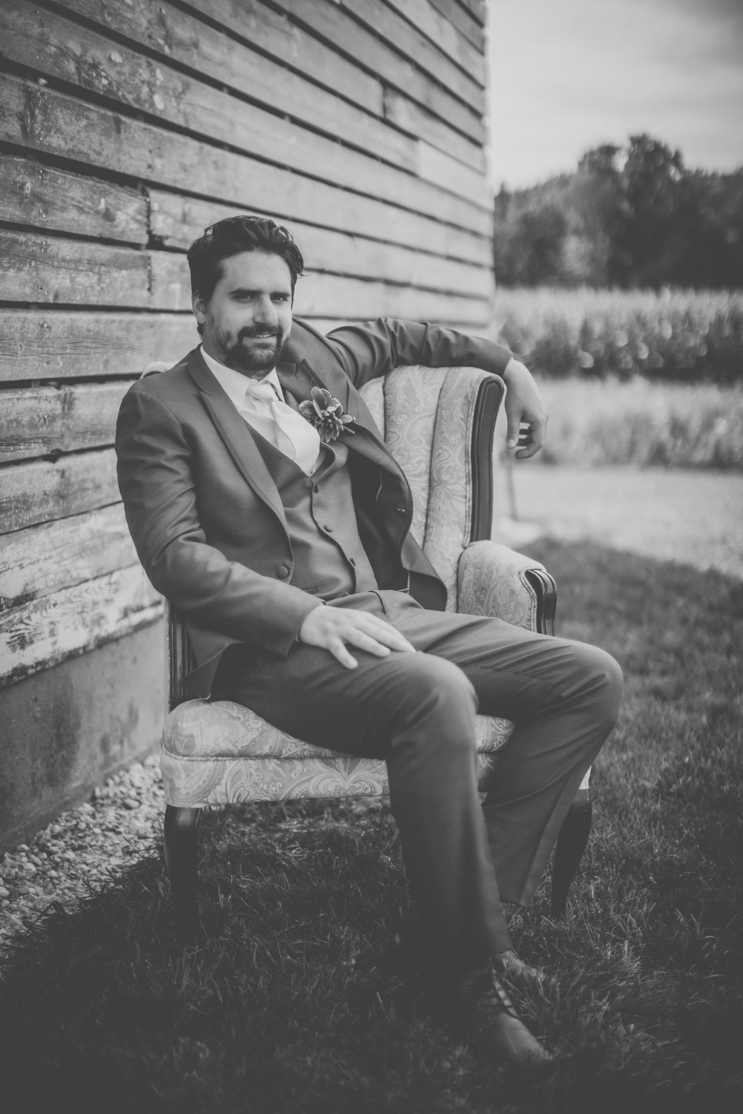 Black and white portrait of a groom in a suit sitting in an elegant vintage chair against a rustic wooden wall.