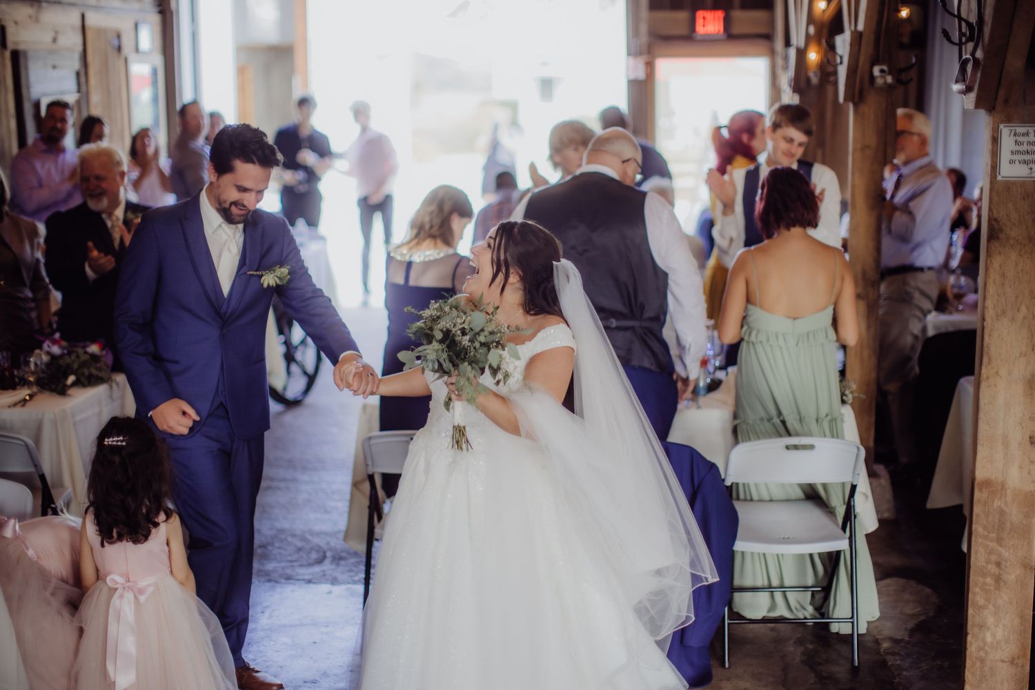 A newlywed couple walks down the aisle at their wedding ceremony surrounded by guests in a rustic indoor venue.