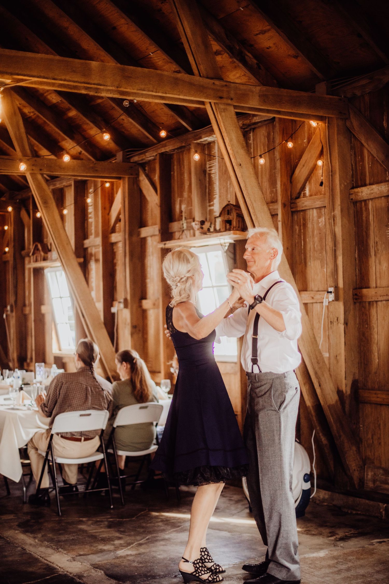 Couples dancing in a rustic wooden barn decorated with string lights and white chairs.
