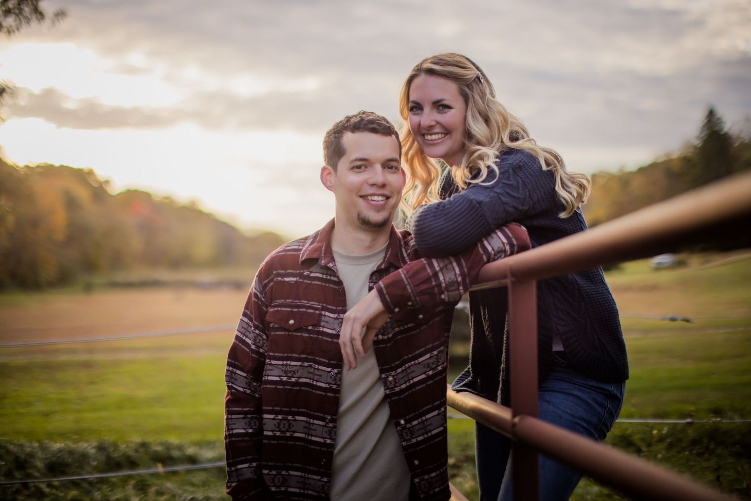 A couple poses together by a wooden fence at sunset in a rustic country setting.