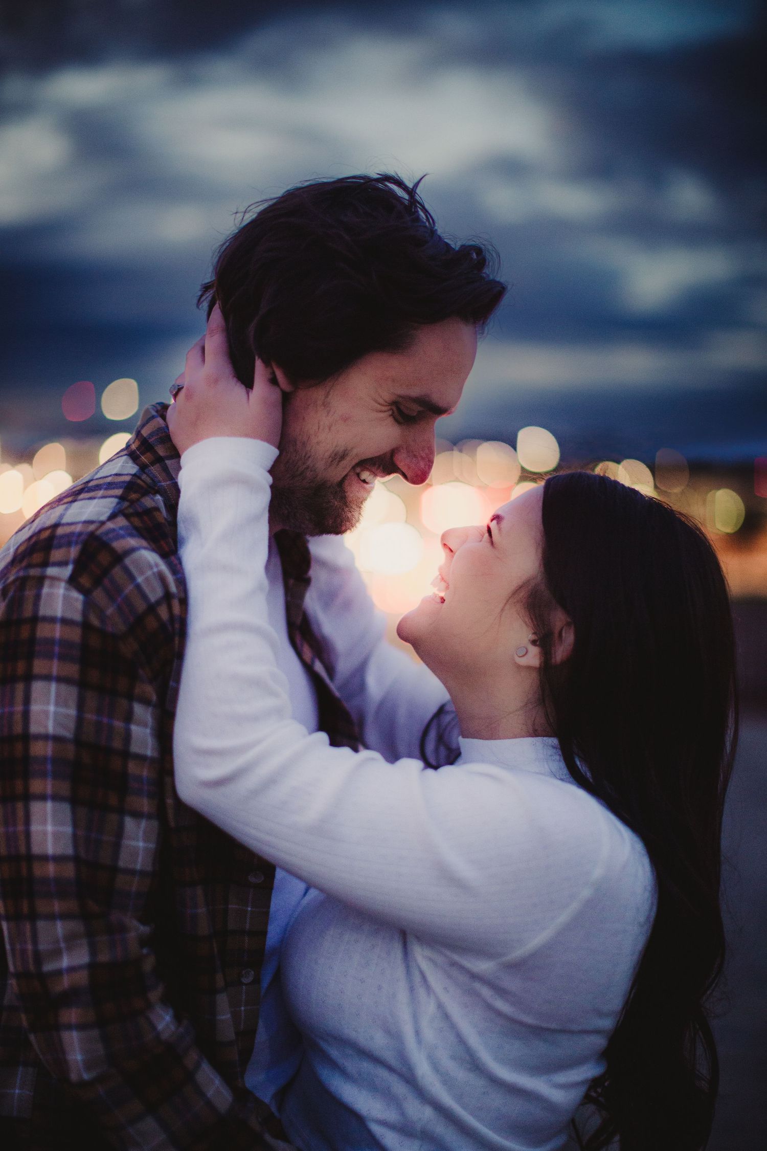 A romantic couple embraces against a sunset backdrop with twinkling lights in the background.