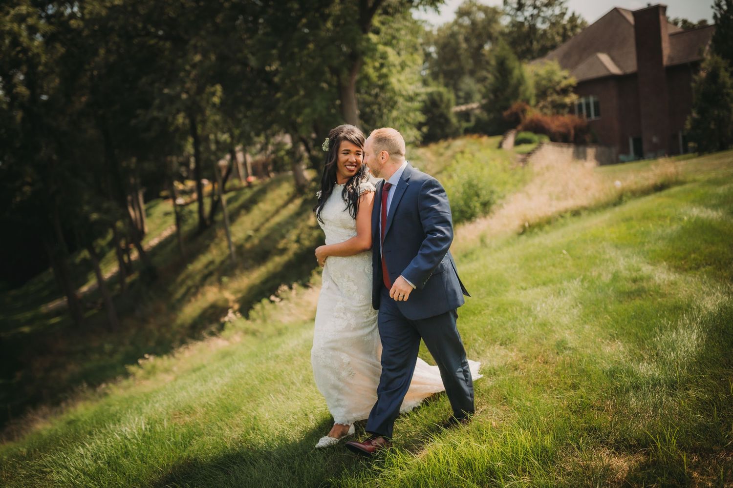 Newlyweds laugh together while walking on a sloped lawn near trees.