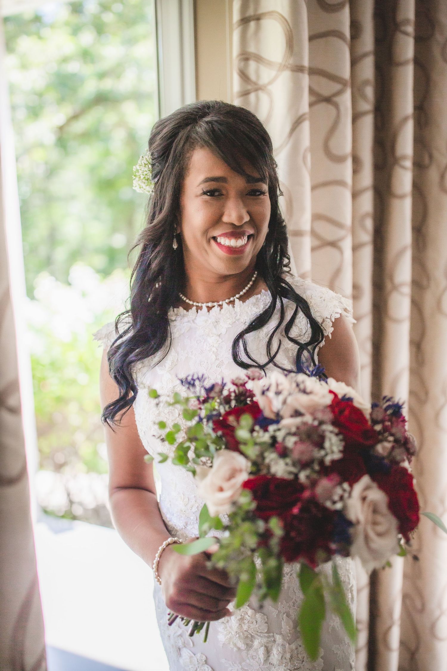 A joyful bride in white lace poses with her burgundy and blush floral bouquet near a window.
