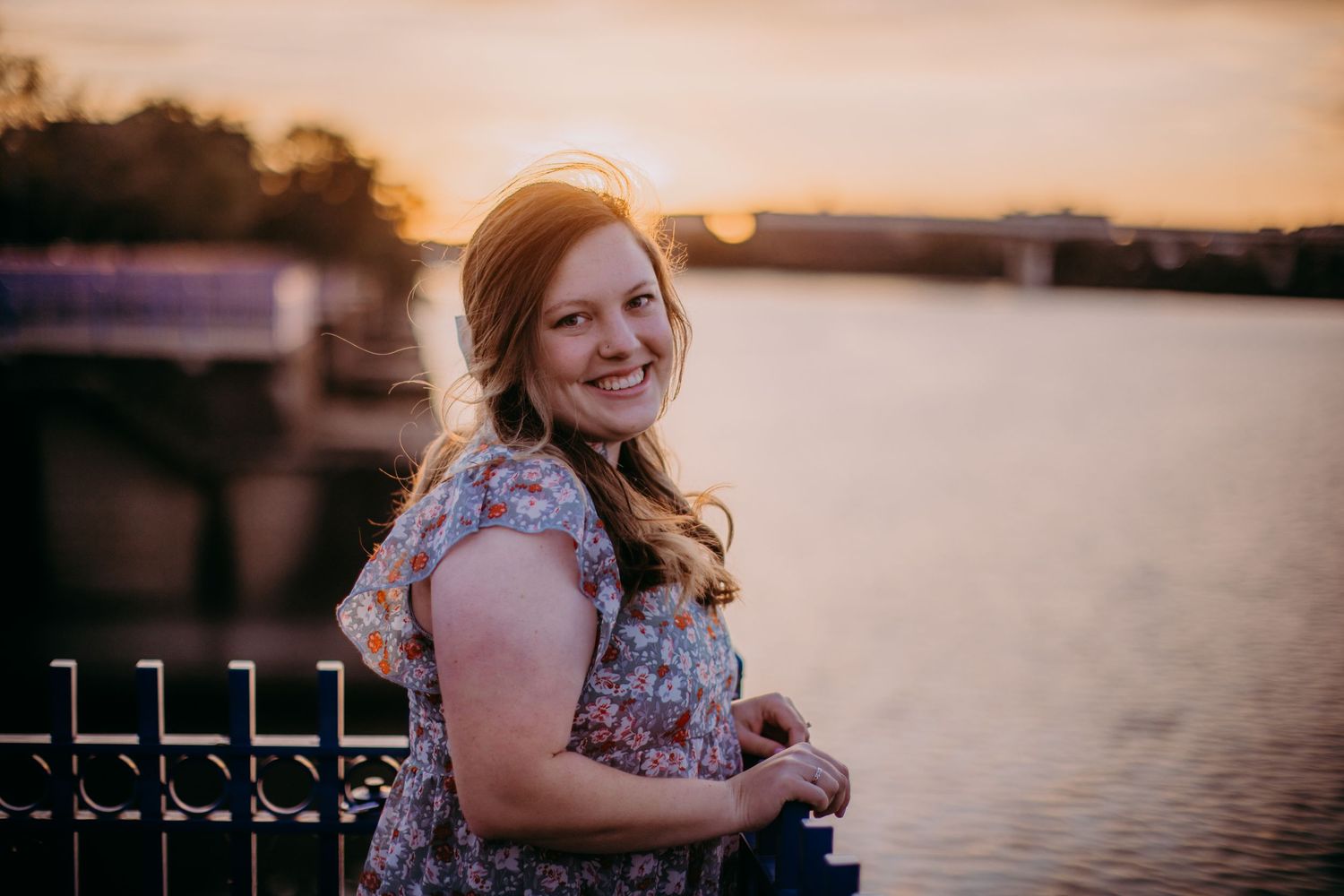 Woman in floral dress poses by metal railing with sunset over water.