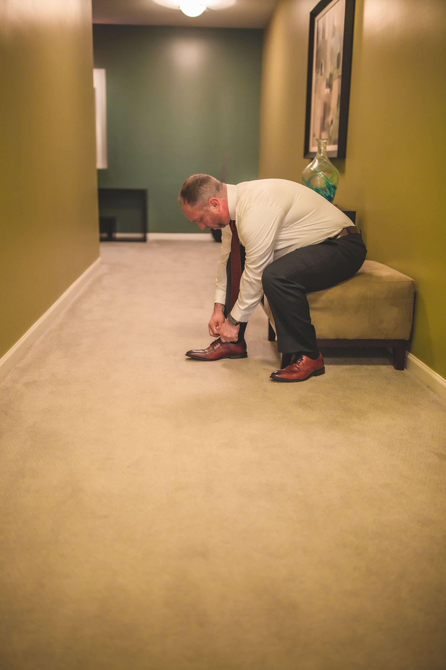 A person adjusts their brown leather shoes while standing in a carpeted hallway with green walls.