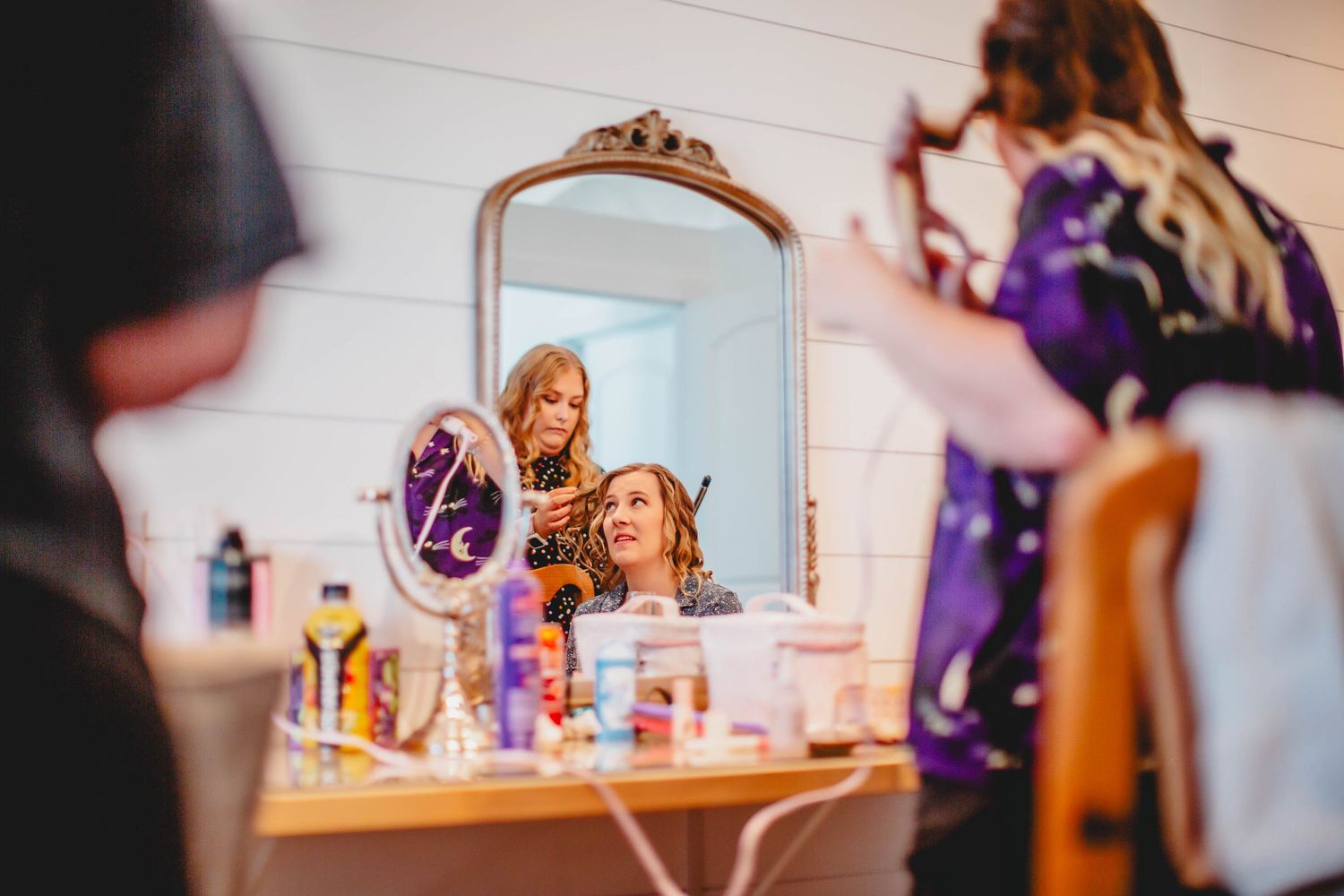 Reflection in ornate mirror shows beauty preparation scene with purple accents.
