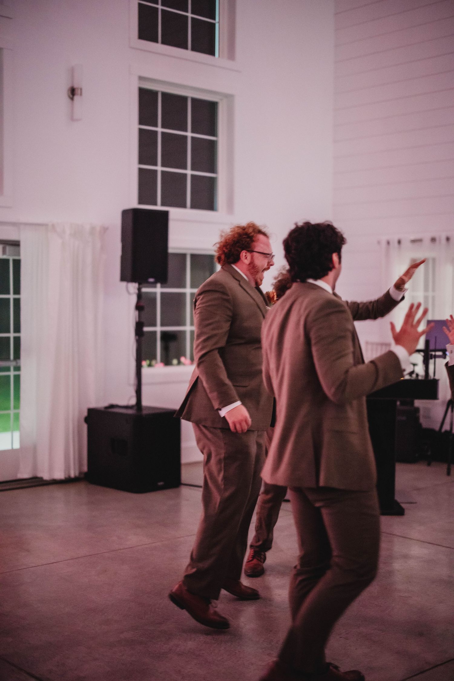 Wedding guests in brown suits dancing enthusiastically on a dance floor with speakers and white curtains.