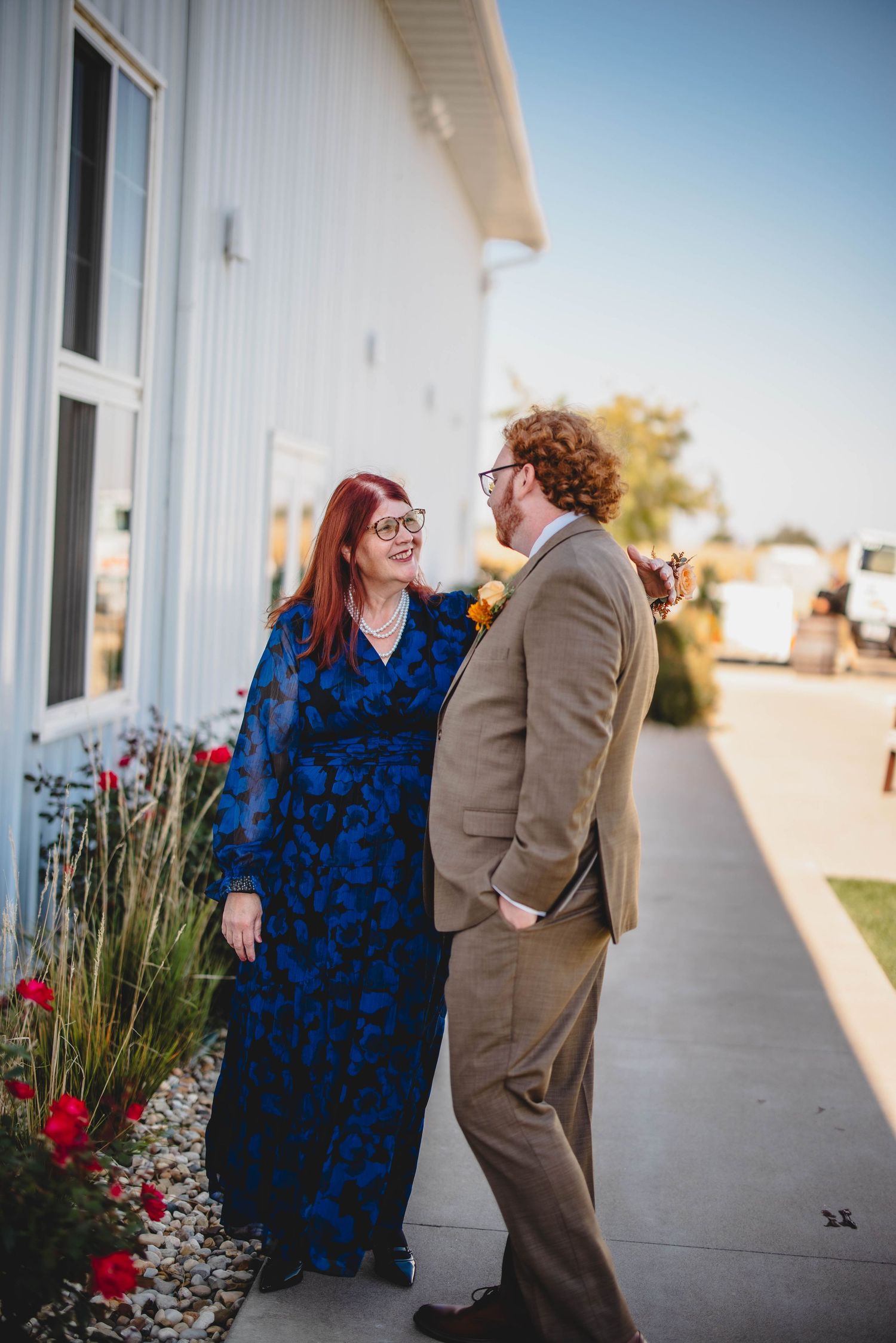 A couple smiling at each other in front of white building with floral landscaping.