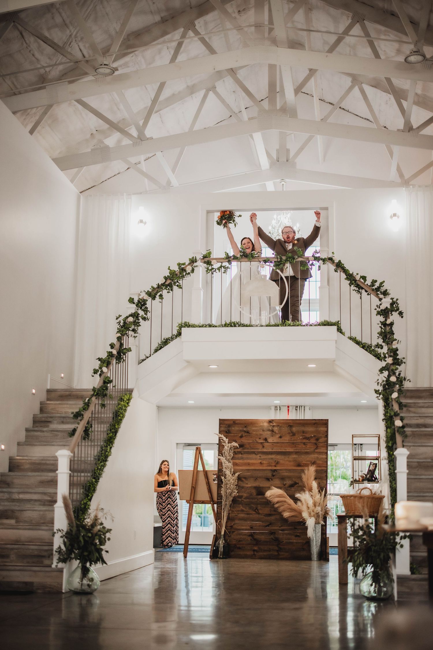 Modern white wedding venue interior with rustic decor features staircase and greenery-adorned balcony.