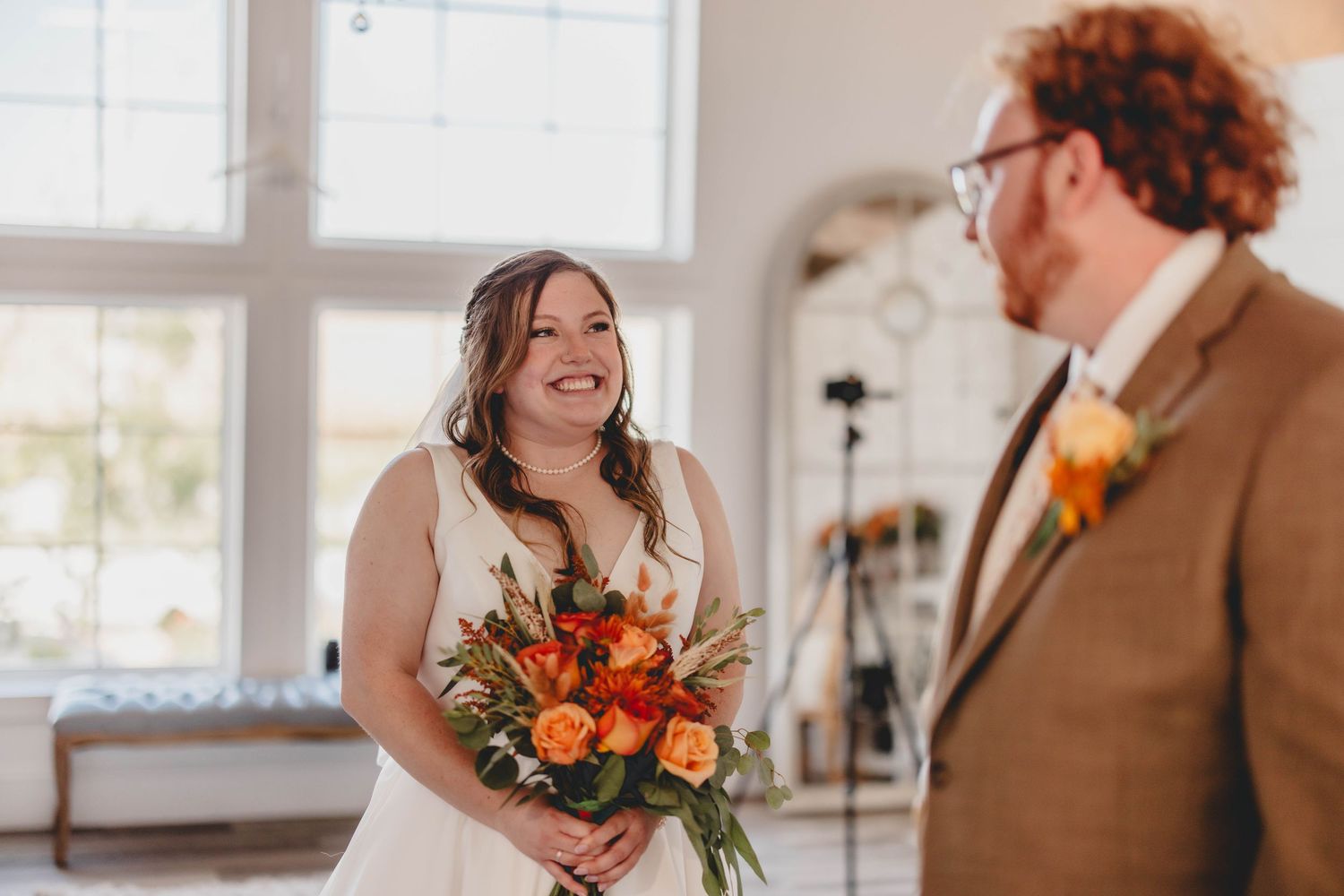 Joyful wedding couple shares emotional first look moment in bright sunlit room with orange floral bouquet.