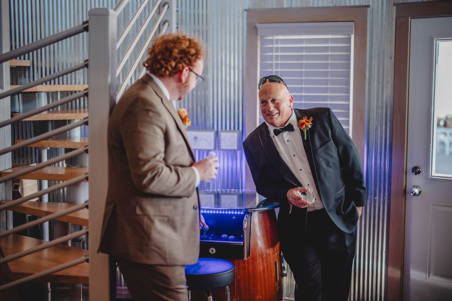 Two men in suits share a laugh while standing near a spiral staircase at an event.