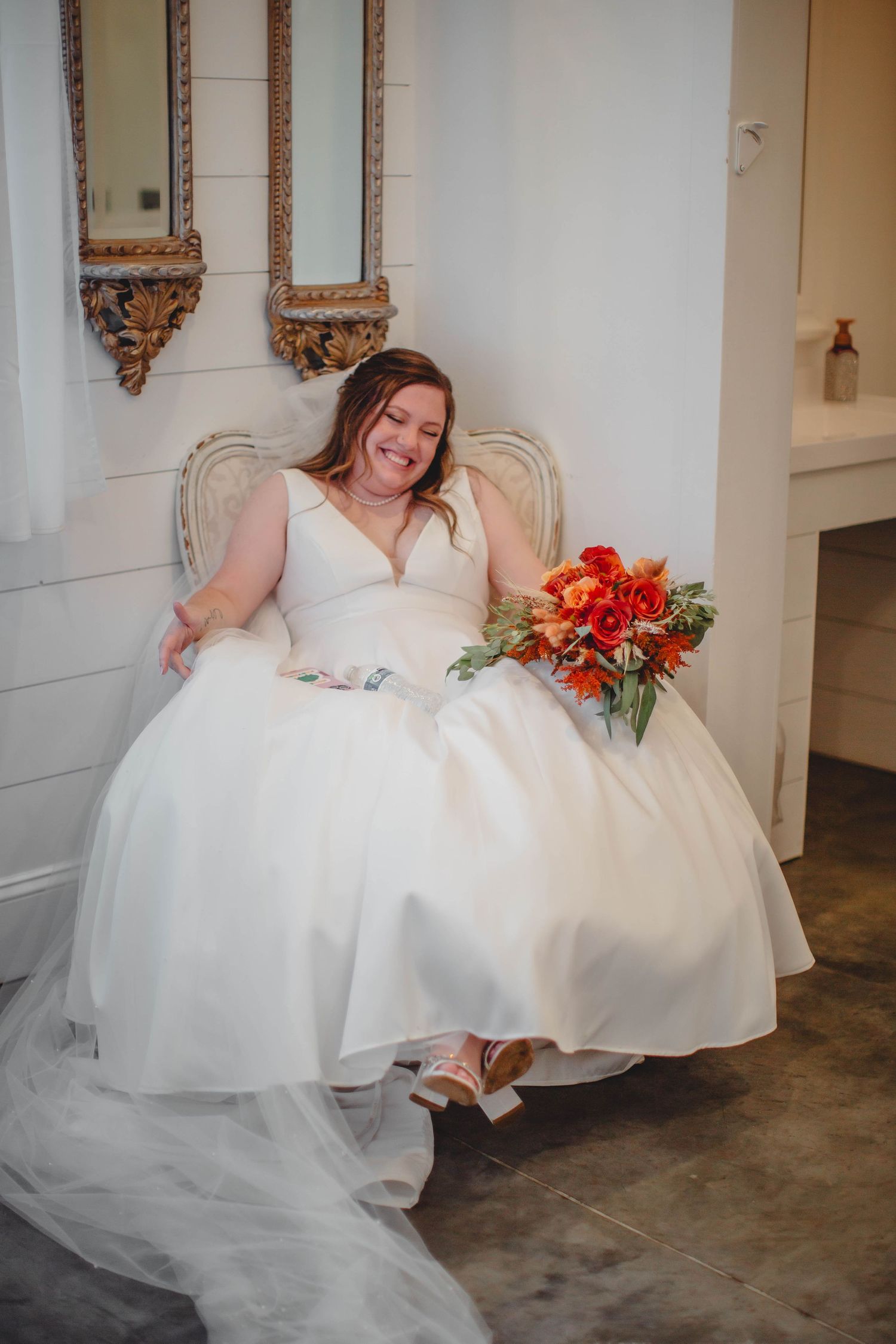 A person in a white wedding gown sits in an elegant chair with a red bouquet.