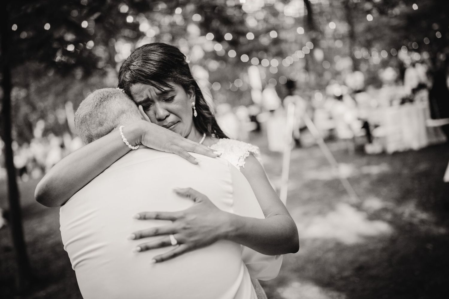 Black and white photo of tender embrace during wedding dance with string lights twinkling in background.