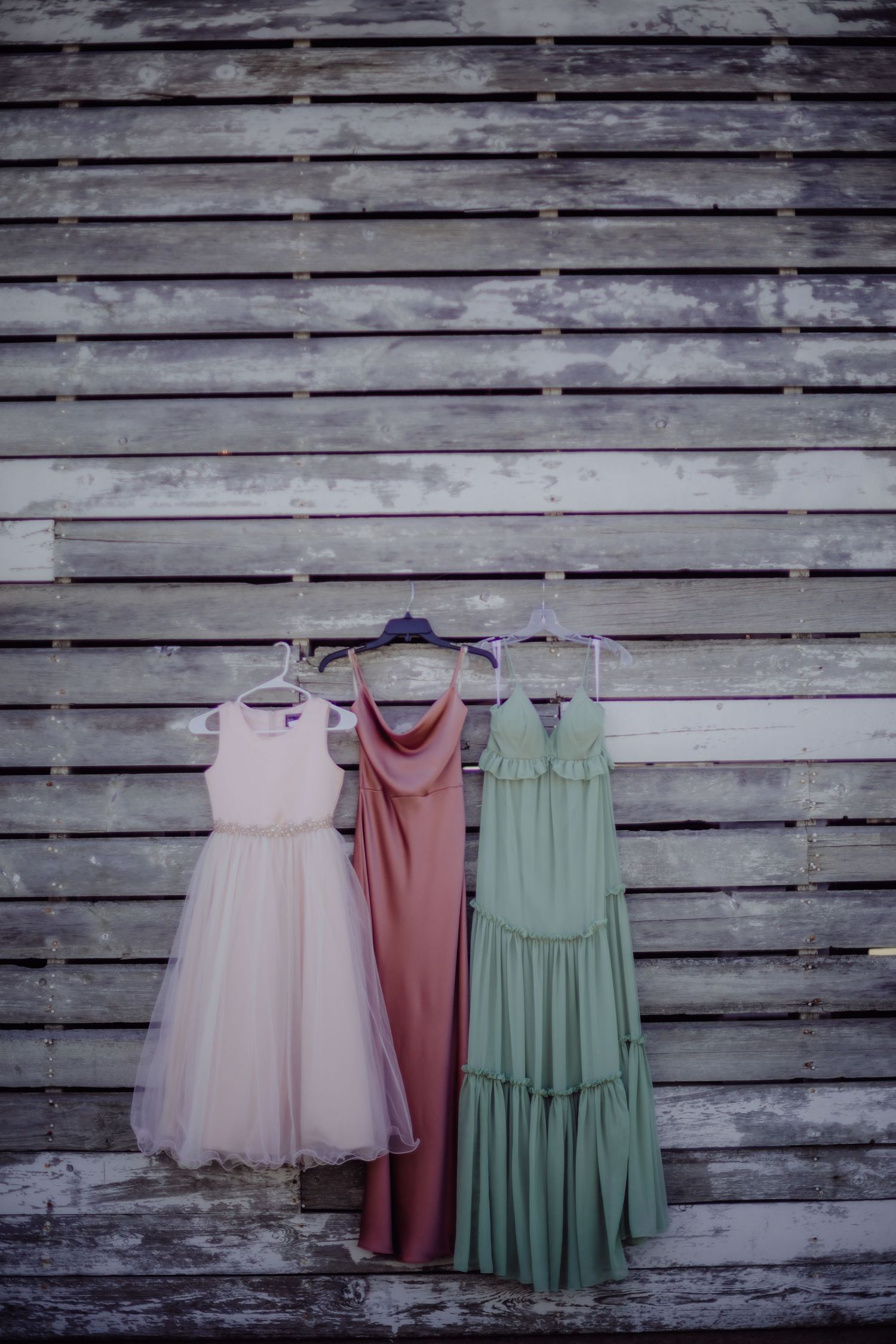Three bridesmaid dresses in pink, blush and sage green hanging against weathered wood wall.