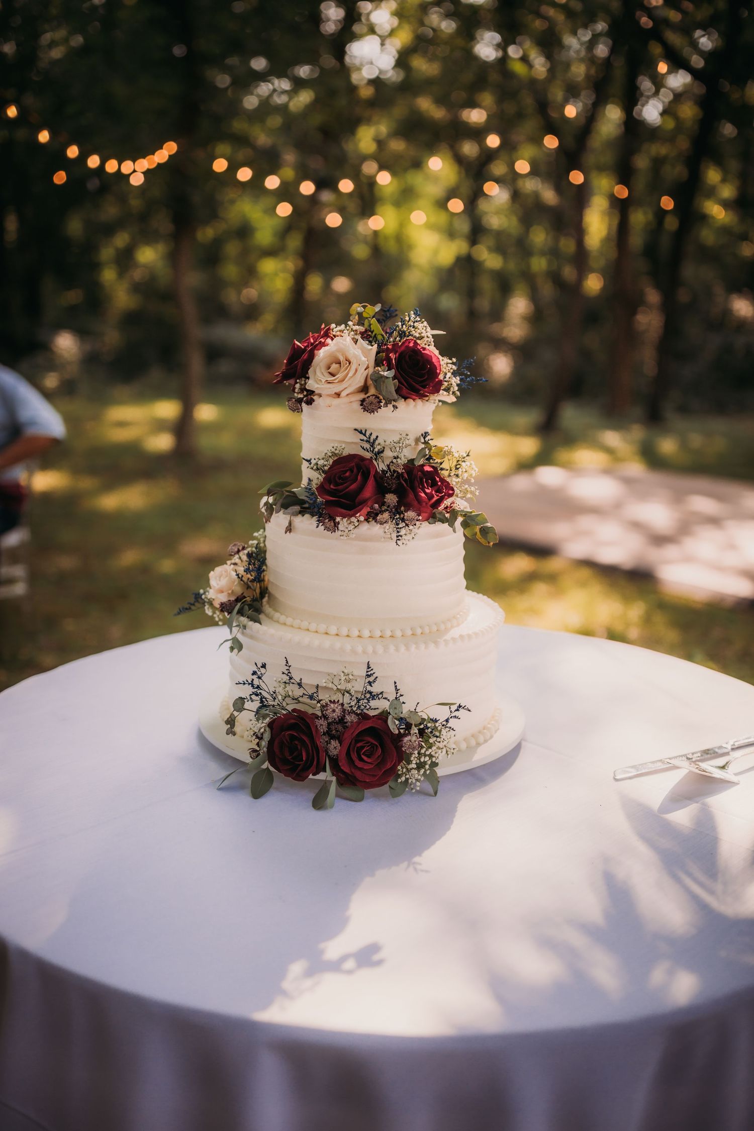 Elegant white wedding cake decorated with burgundy roses and delicate floral accents on a white tablecloth.