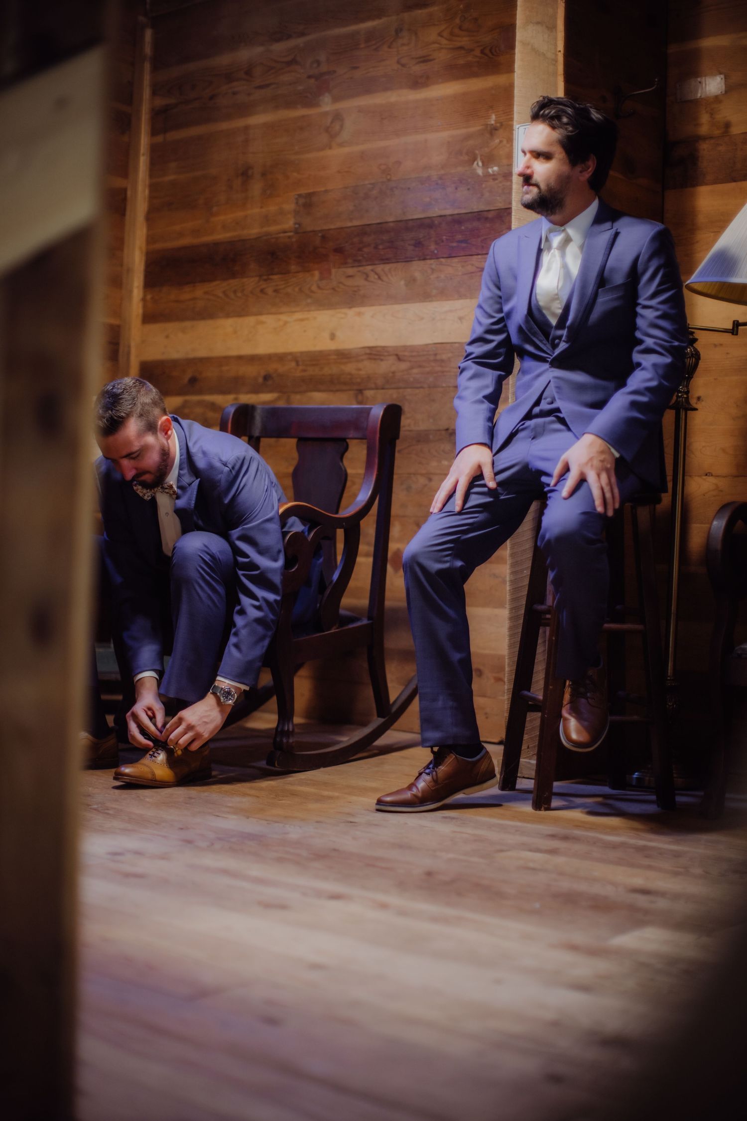 Two men in navy suits getting ready while sitting on wooden chairs.