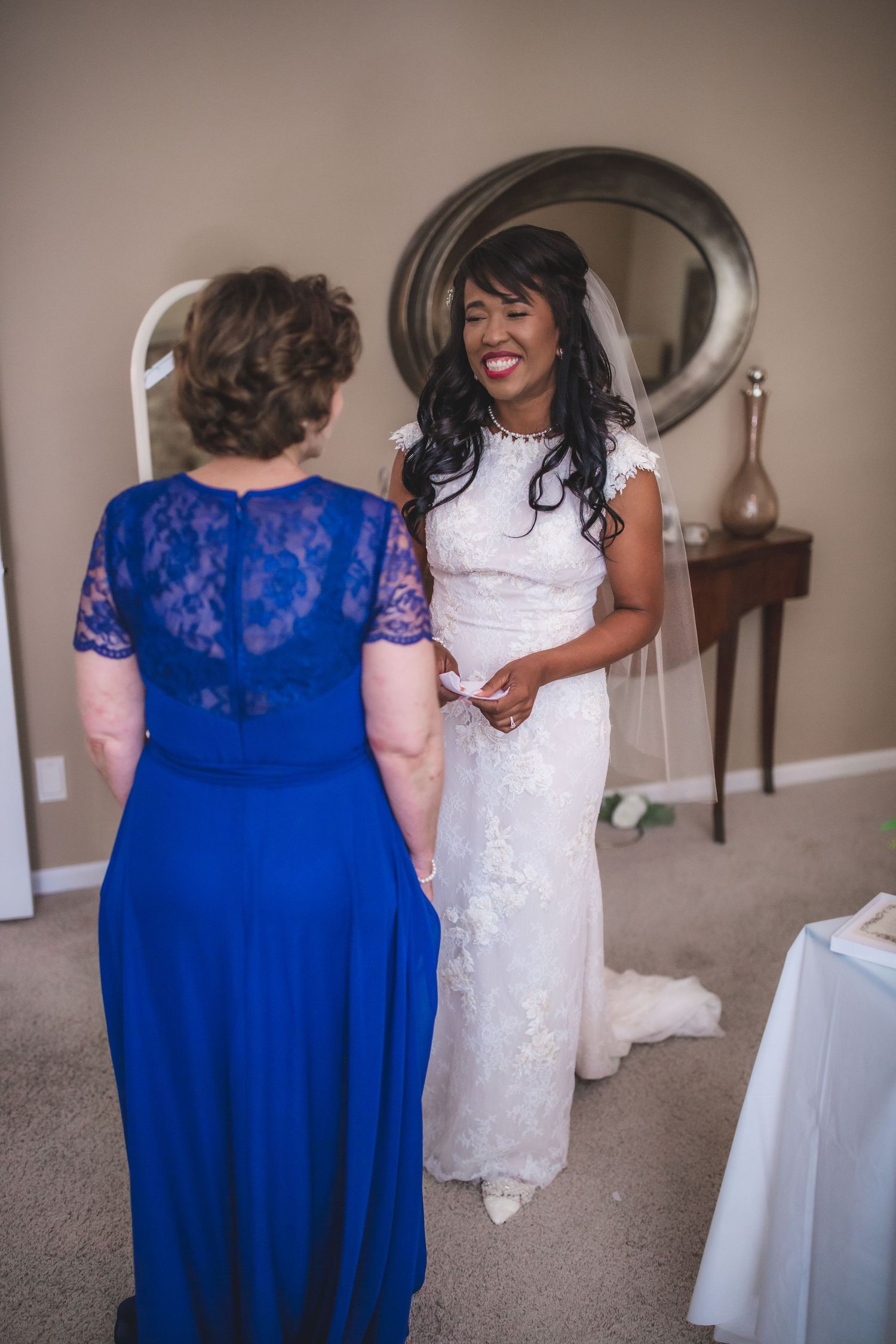 A bride in white lace shares a joyful moment with a woman in a royal blue dress.