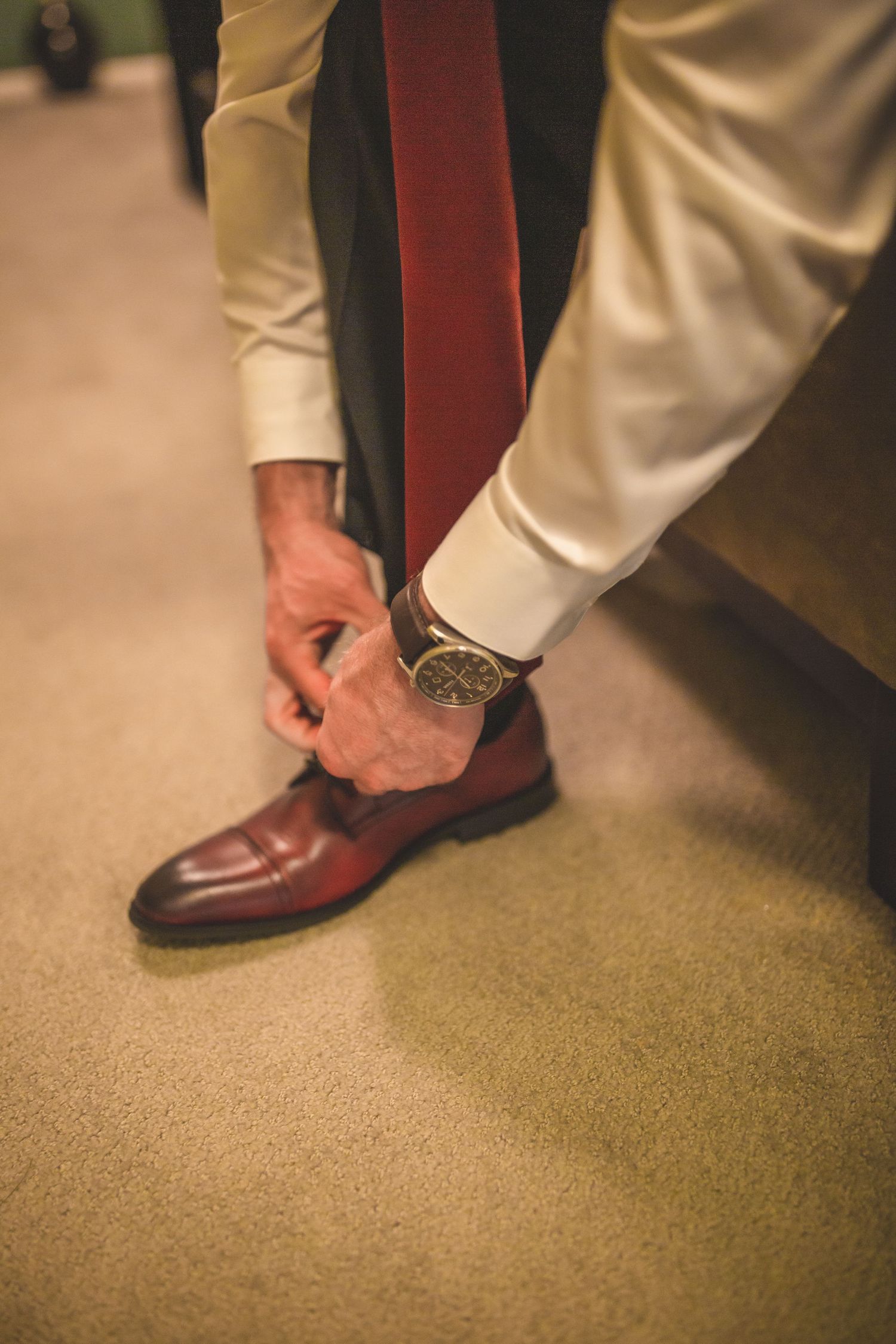 Close-up shot of hands adjusting a brown leather dress shoe with white shirt sleeve visible.
