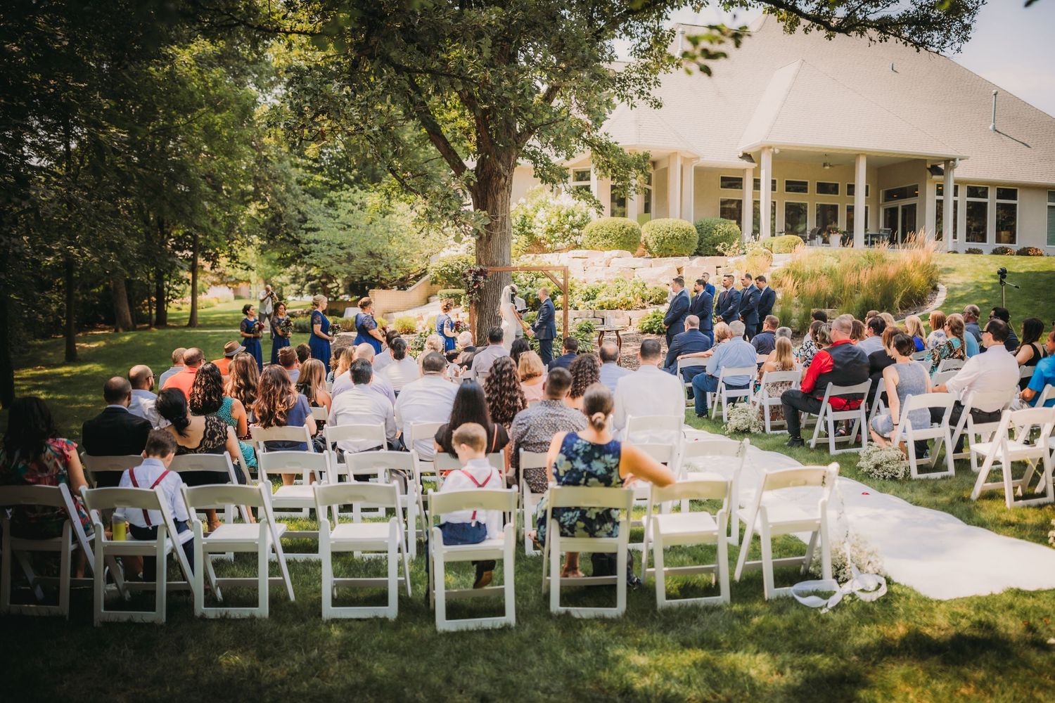 Wide view of a garden wedding ceremony with white chairs arranged in rows on a sunny afternoon.