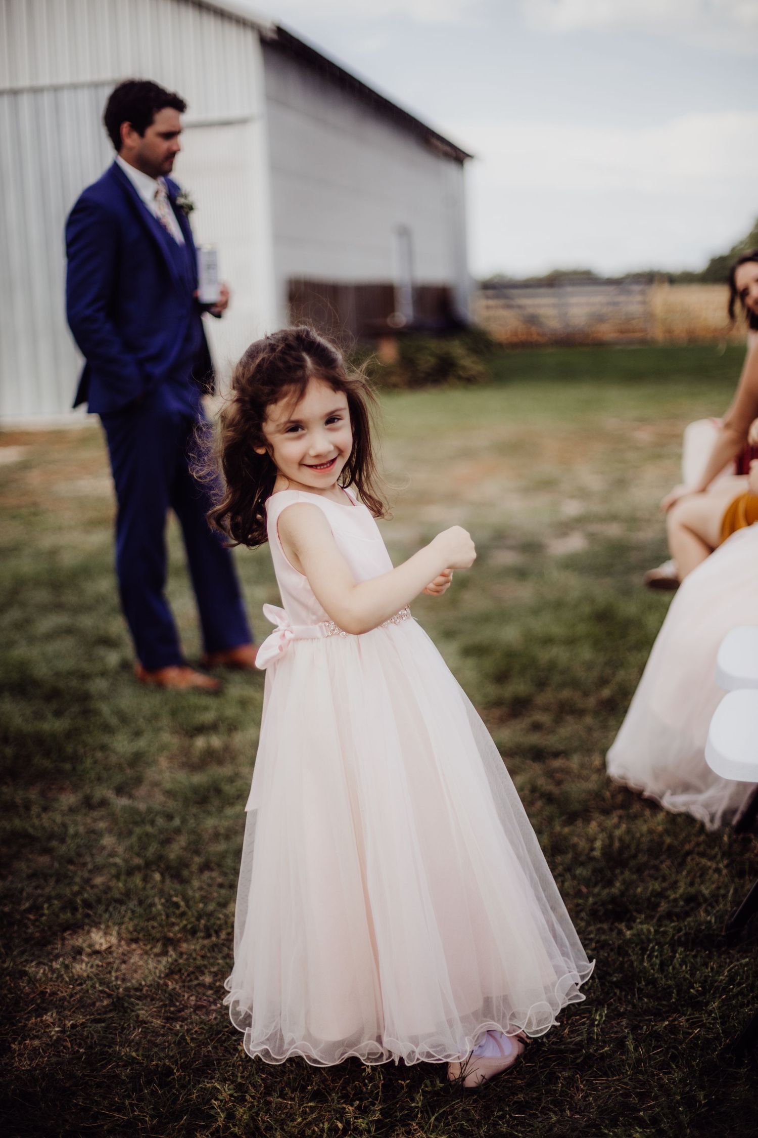 A young flower girl in a pink tulle dress twirls joyfully on the grass outside.