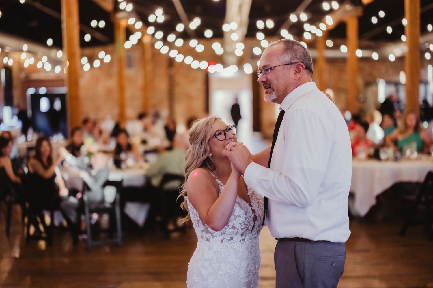 Tender moment shared during wedding dance in rustic reception hall with string lights.