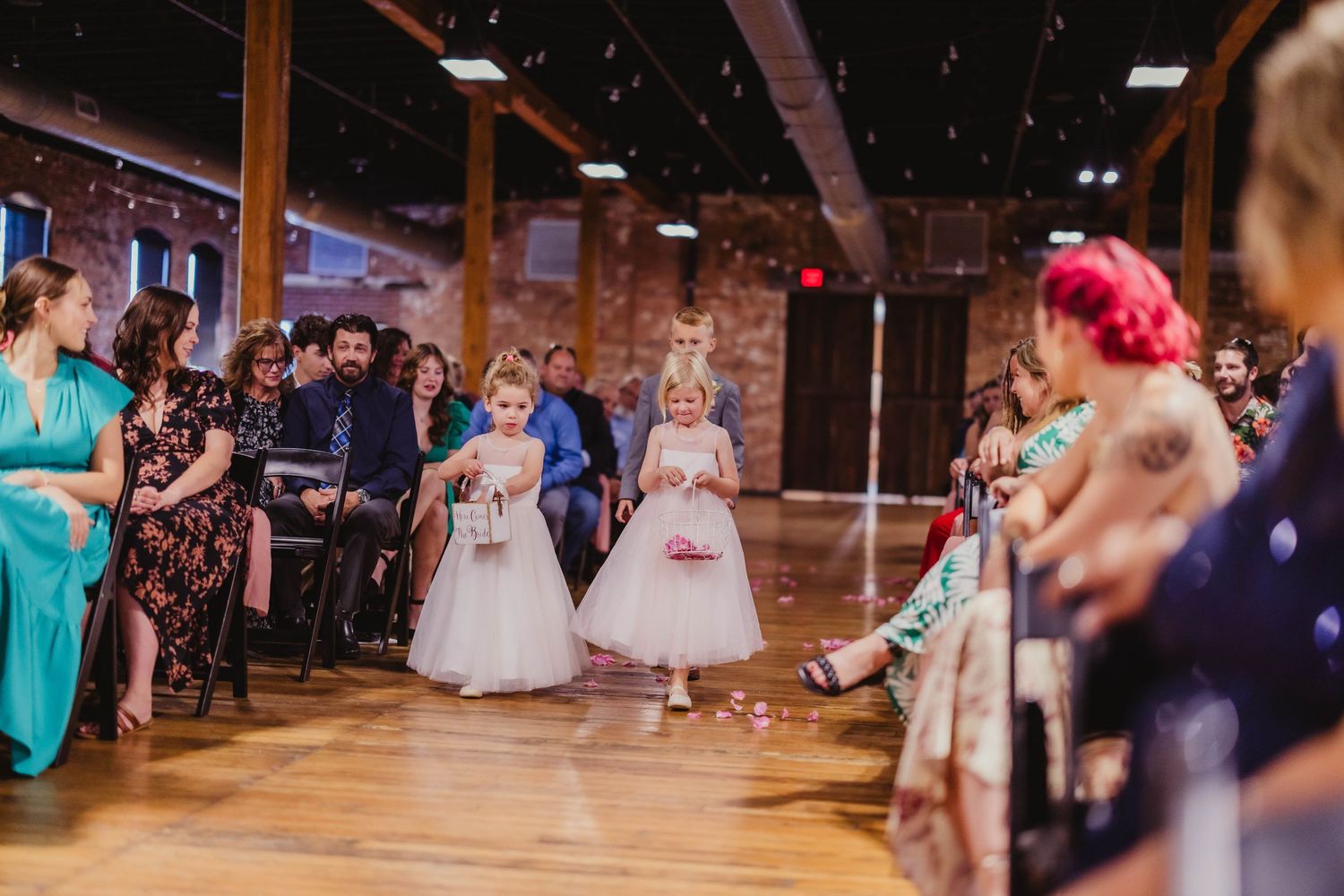 Two flower girls in white dresses walk down the aisle during a wedding ceremony.
