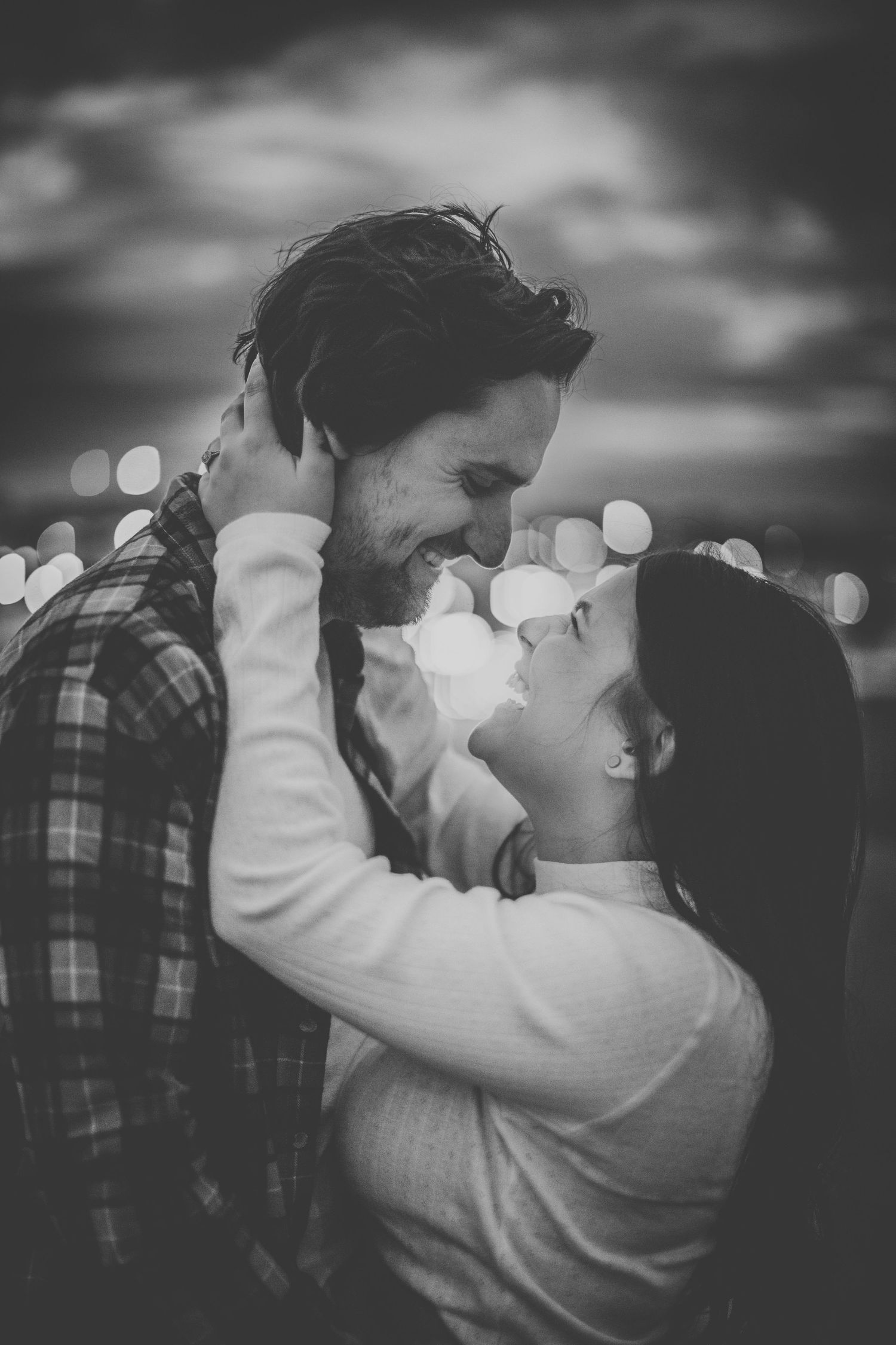 A couple laughs together in black and white with twinkling city lights in the background.