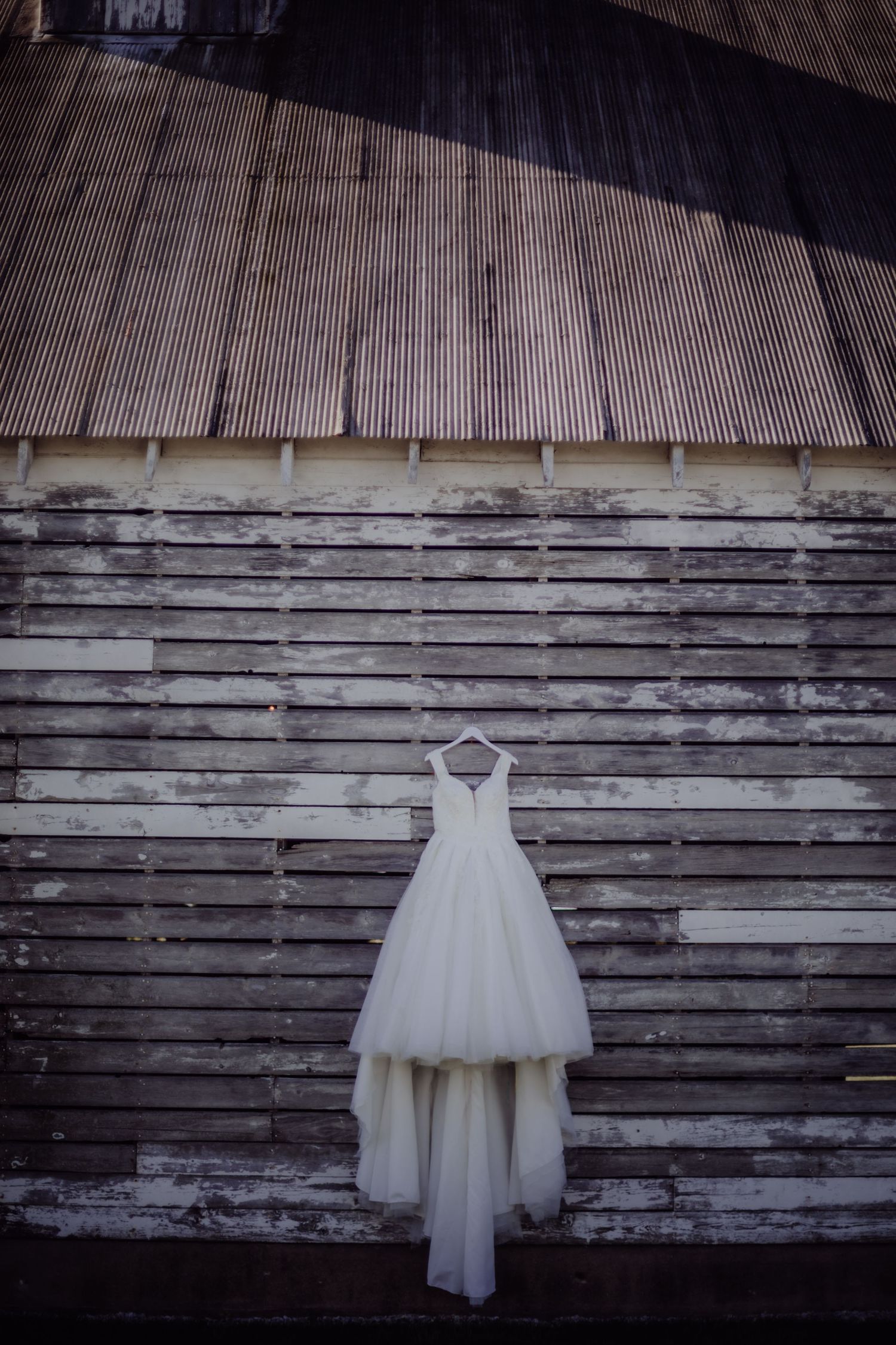 White wedding gown hanging on rustic wooden barn wall with corrugated metal roof.