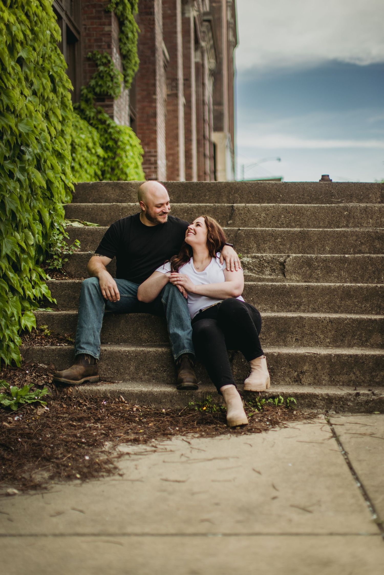A couple relaxes on concrete steps beside an ivy-covered brick wall in casual outdoor photo session.