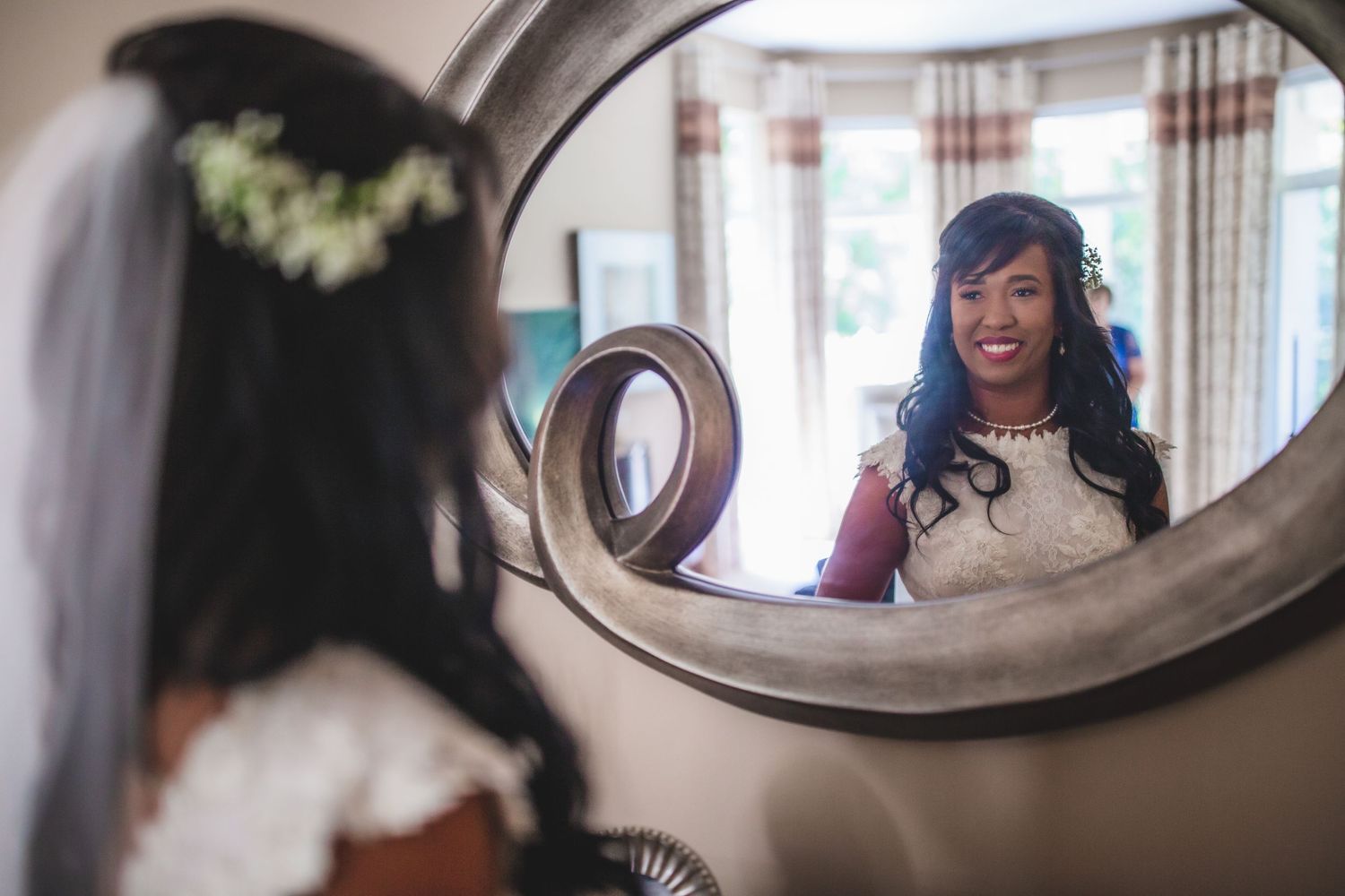 A joyful bride looks at her reflection in a vintage oval mirror during wedding preparations.
