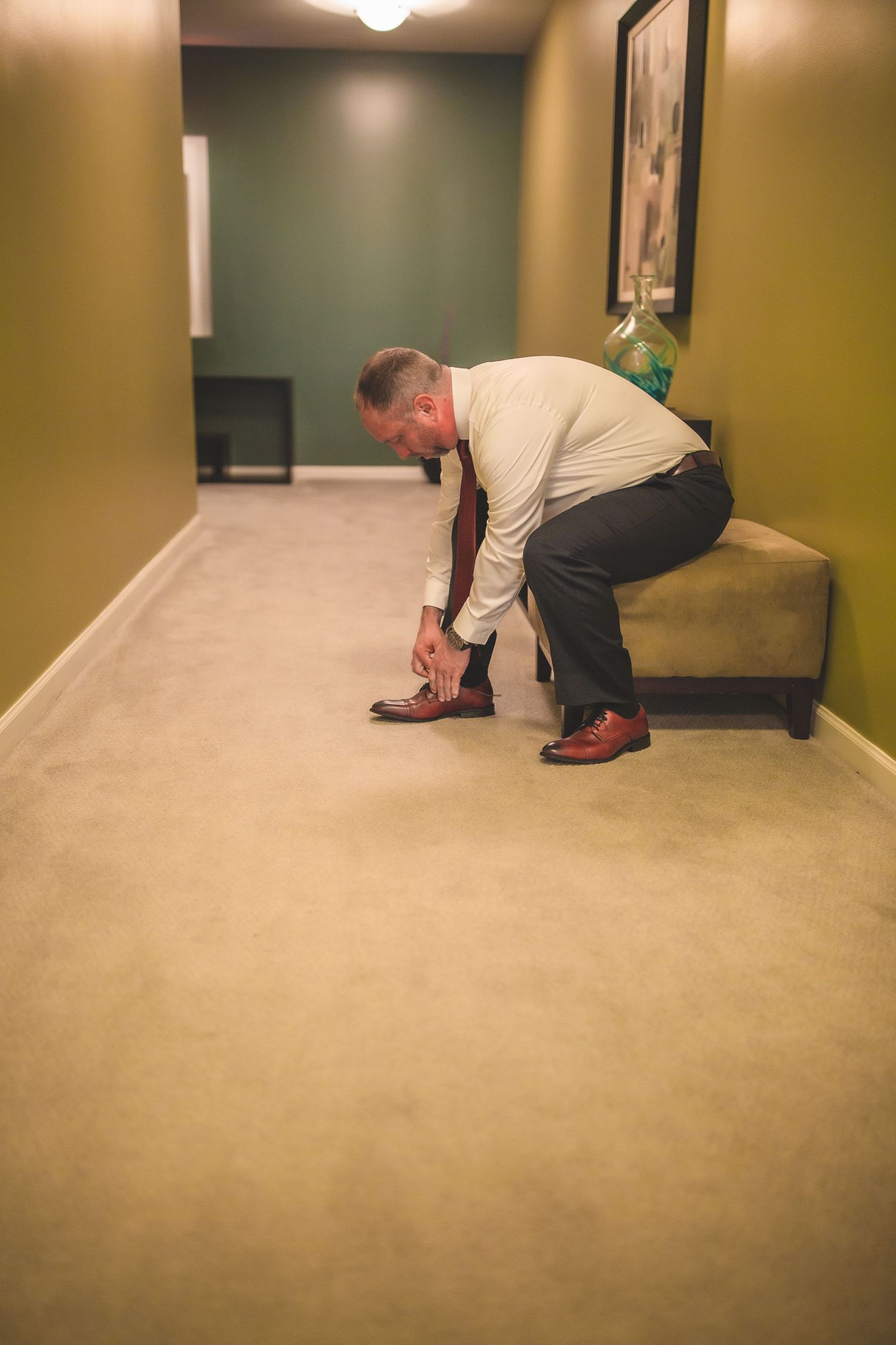 A person in formal wear bends down to adjust their brown dress shoes in a hallway with beige carpet.