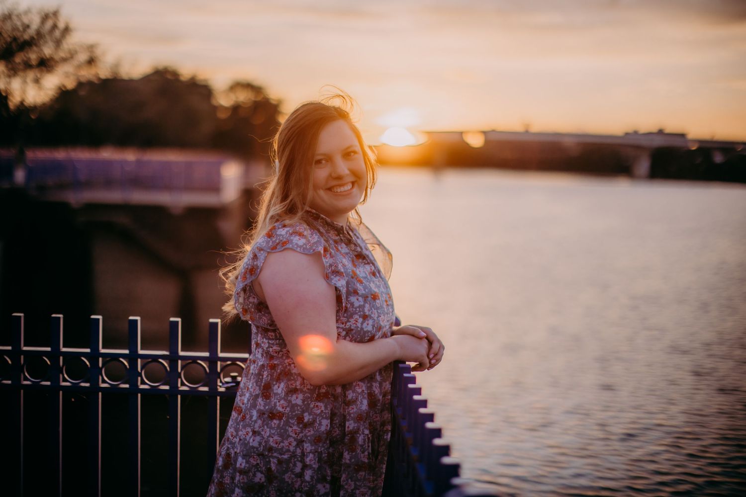 Woman in floral dress smiles by waterfront railing during golden hour.
