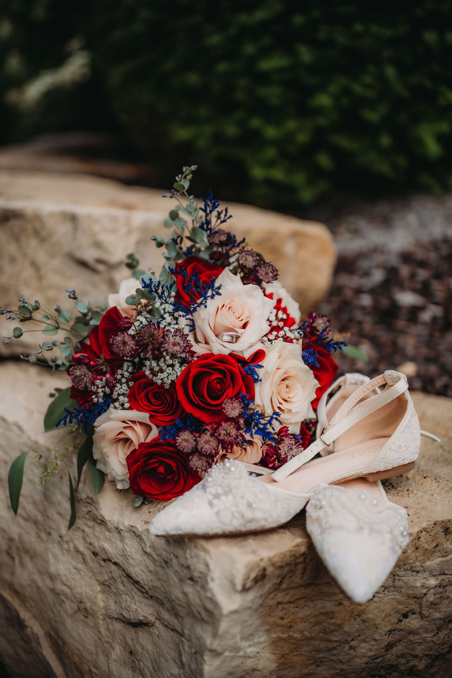Wedding bouquet in red, white and blue tones paired with lace bridal shoes on rustic stone.