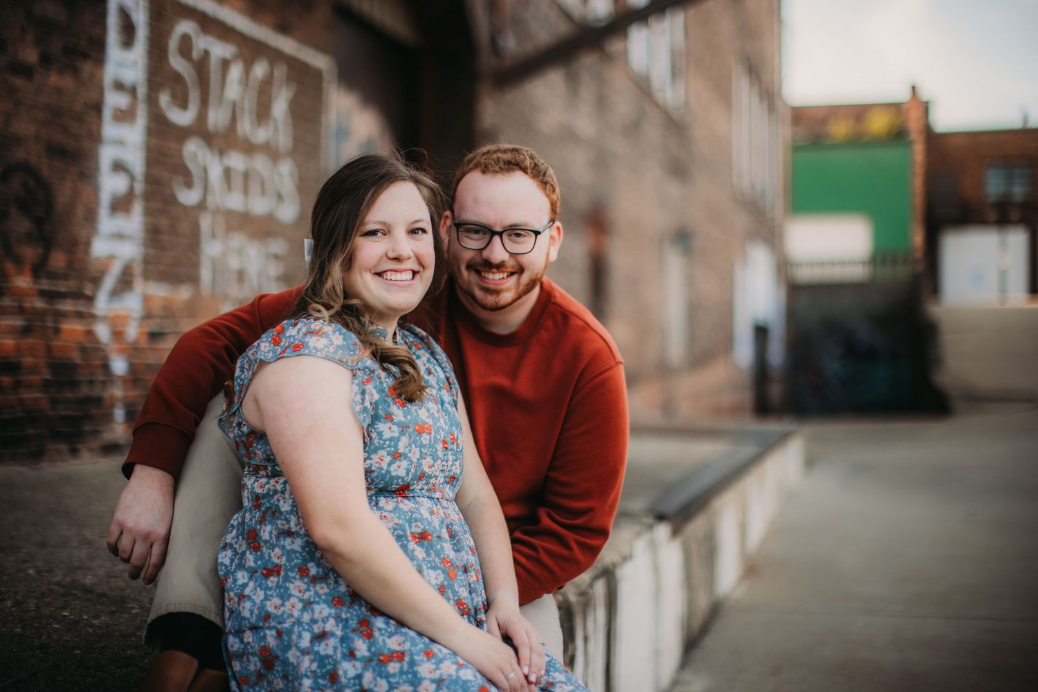 A romantic couple sits together against an industrial brick backdrop.