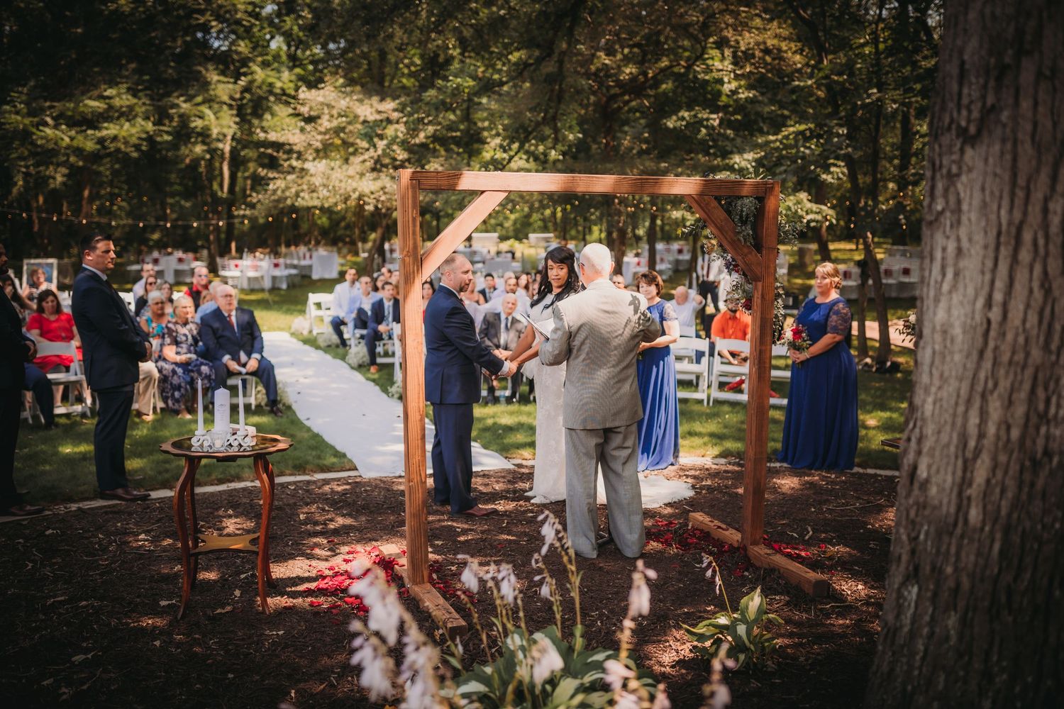 Wedding guests gather for an intimate outdoor ceremony surrounded by mature trees and natural shade.